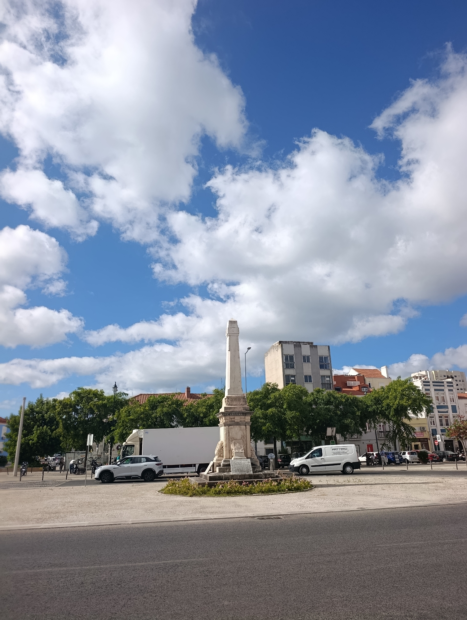 This is a vertical, eye-level, full-shot photograph of a sunny day in Setúbal, Portugal, looking across a paved area towards buildings and trees. In the foreground, a gray asphalt road is visible. Beyond this, a light-colored paved area contains a monument, trees, and parked vehicles. The monument is a tall, white, stone structure with intricate carvings and a pedestal, topped by a square obelisk. To the left of the monument, a white SUV is parked. Behind the SUV, a large white truck is parked. To the right of the monument, a white van is parked, with text on its side that appears to read "PRIME TECH" and "SOLUCOES INTEGRADAS".
Further back, a line of lush green trees forms a backdrop. Behind the trees, several buildings are visible. The closest building to the left appears to be a light blue residential building. In the center, a tall, gray, multi-story building with many windows is prominent. To the right of this, a building with red-tiled roofs is visible, followed by more multi-story residential buildings with white facades and blue-trimmed windows.
The sky is a vibrant blue, dotted with fluffy white clouds, suggesting fair weather. The scene captures a street or plaza area in an urban environment, likely a public square, given the monument and the open paved space.