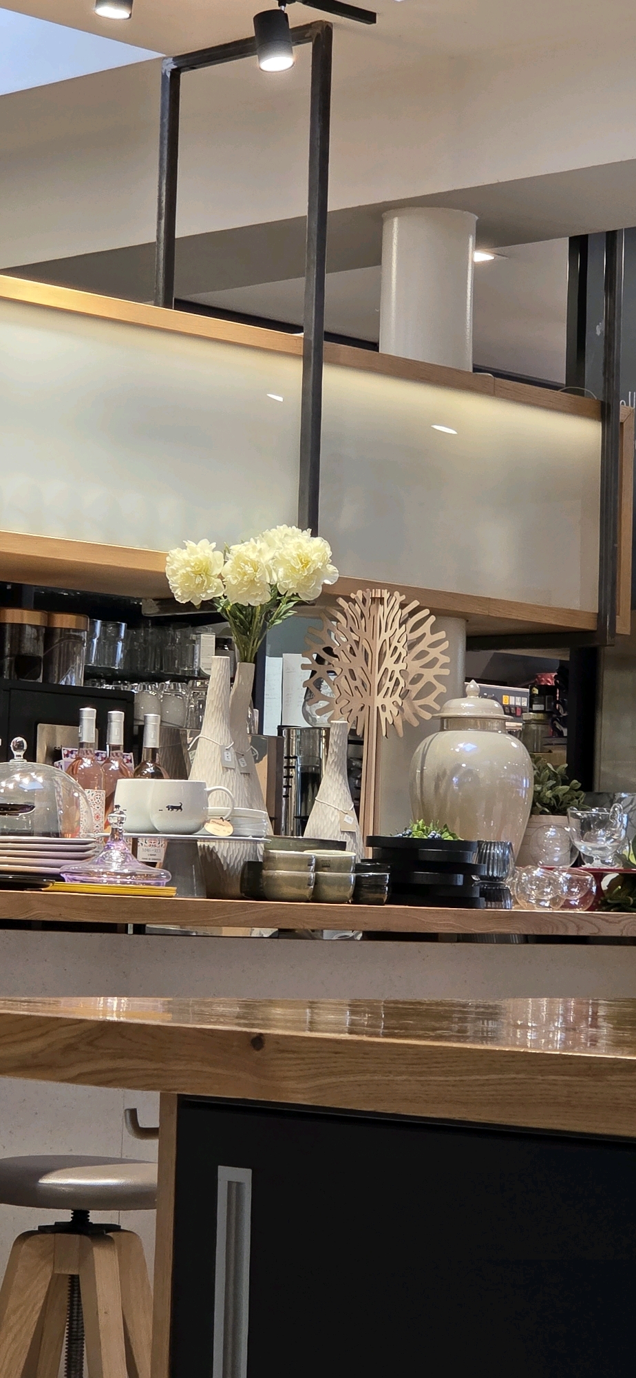 The image depicts a shelf display in a store located in Kehl, Germany. The foreground features a wooden counter with a reflective surface. The shelf itself is made of wood and is populated with various decorative and functional items.
Prominently displayed are a bouquet of yellow flowers in a white vase, two textured white vases, and a decorative wooden tree sculpture. To the right, a large, cream-colored ceramic jar with a lid sits on a black platter. A stack of smaller bowls and cups are also visible, some with a metallic sheen. In the background, there are shelves filled with glassware, bottles (including what appear to be rose wine bottles), and other assorted items. The overall lighting suggests an indoor environment, possibly a home goods store or a cafe with a retail section. The lighting fixtures are visible at the top of the image, attached to dark metal framing. The presence of a stool in the lower left corner suggests this is an area accessible to customers or staff.