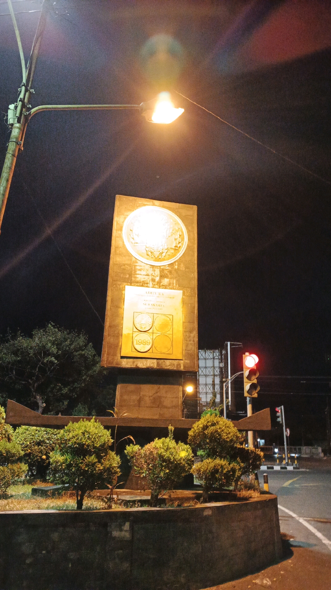 The image is a night-time photograph of a monument in Surakarta, Indonesia. The monument is a tall, rectangular structure with a circular emblem at the top and a plaque below it. The plaque has text and four circular images, with the year "1989" prominently displayed. The monument is illuminated by a street light, which casts a warm glow on its surface and creates lens flares in the sky.
In the foreground, there are several small, neatly trimmed bushes arranged in a planter. Behind the monument, a traffic light is visible, with its red light illuminated. The surrounding environment appears to be an urban setting, with a road and some buildings in the background, including a large structure with a lattice framework. The sky is dark, indicating it is nighttime. The image seems to capture a significant landmark within the city.