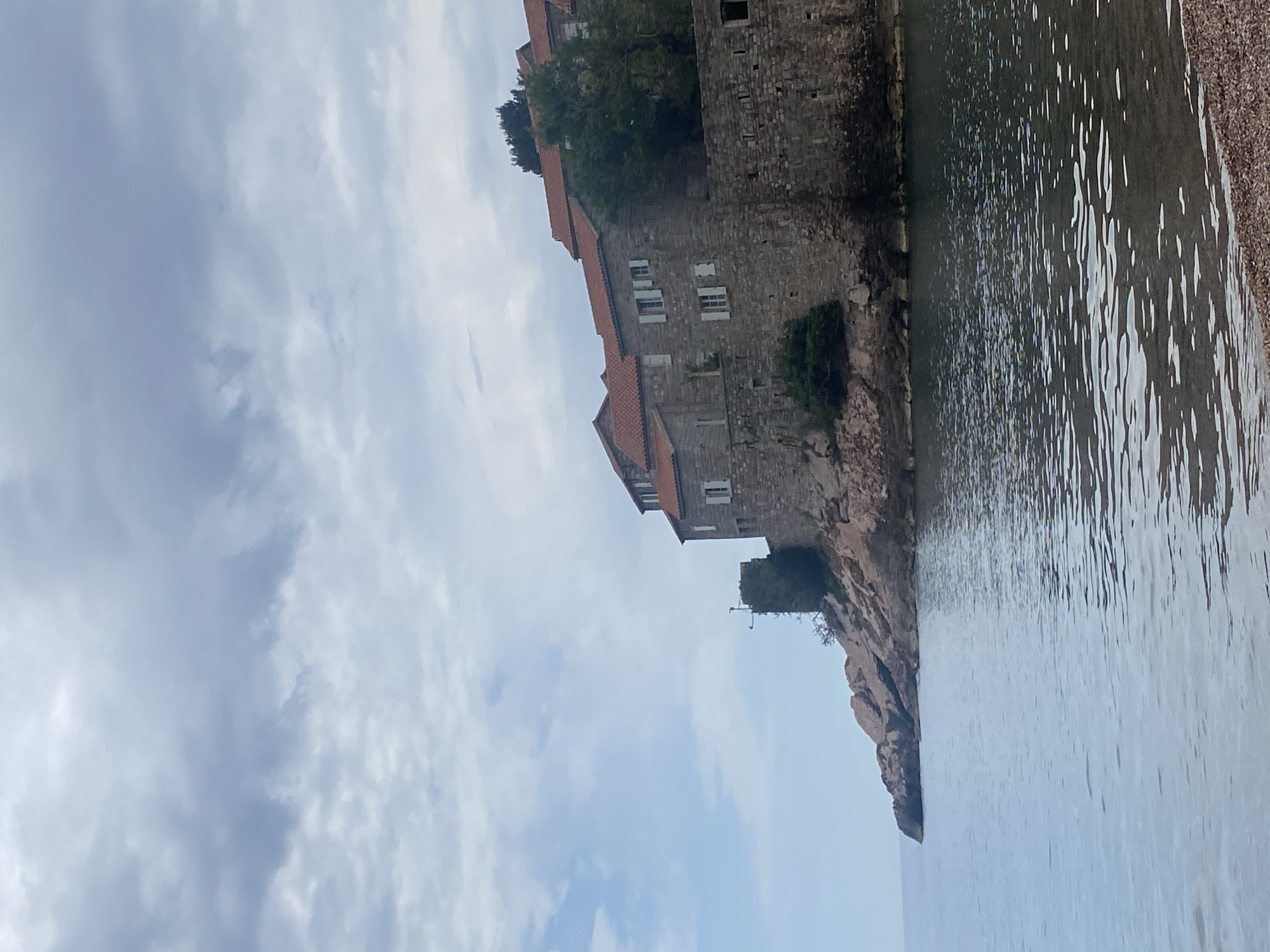 The image depicts Sveti Stefan, Montenegro. The stone buildings with red tile roofs are situated on a rocky promontory extending into the sea. The water is calm, reflecting light. The sky is overcast with a cloud-filled sky. The buildings consist of multiple levels and windows. Some vegetation is visible on the rocks and around the buildings. A beach is barely seen.