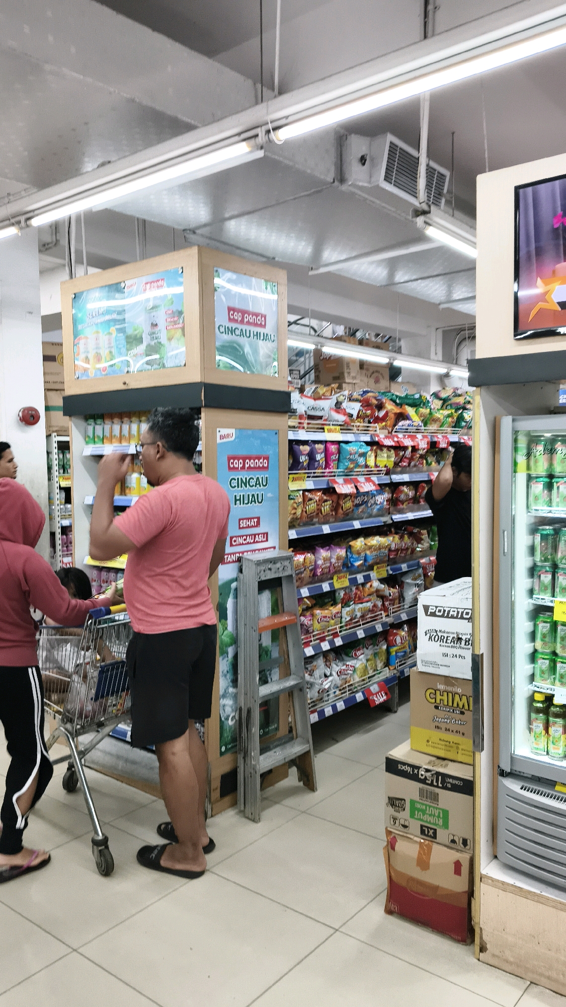The image depicts the interior of a supermarket in West Jakarta, Indonesia. In the foreground, a man wearing a pink t-shirt and black shorts stands facing left, with his hand near his mouth. He is positioned next to a shopping cart with a person in a pink hoodie and black pants standing in front of it. To the right of the man, a metal ladder leans against a display shelf filled with various packaged snacks. Further into the background, more shelves are stocked with a wide assortment of food items. A refrigerator unit is visible on the far right, containing bottled drinks. The overall environment suggests a typical retail setting with fluorescent lighting and tiled flooring.