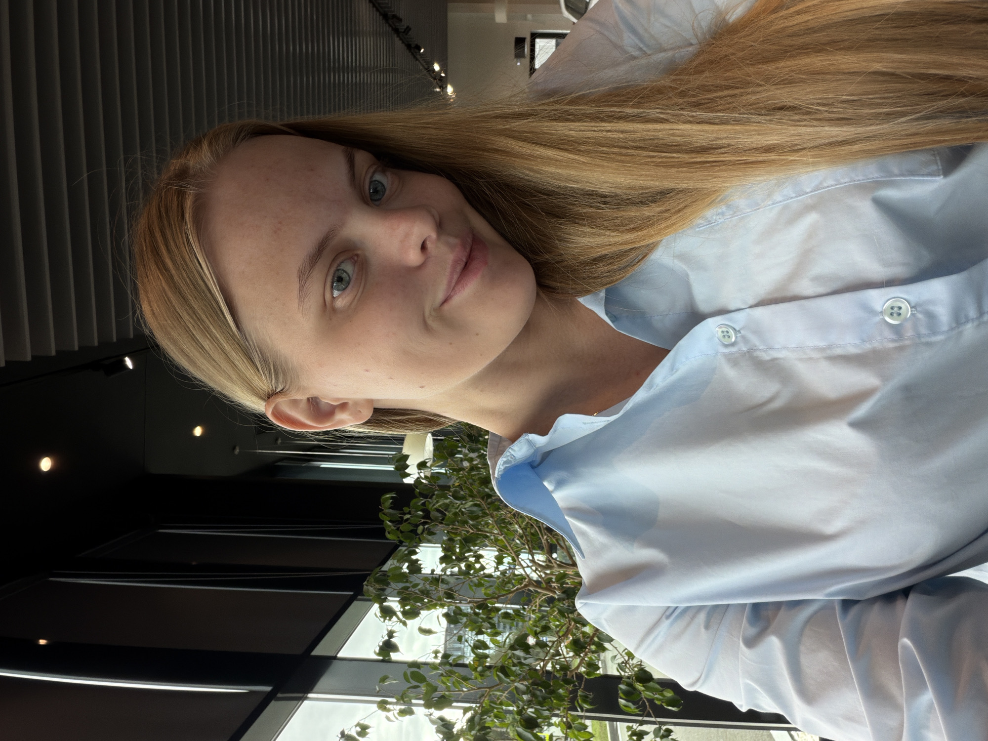 A light-skinned woman with long blonde hair, wearing a light blue button-down shirt, is the primary subject of the image. She is indoors, near a window with a view of a green leafy plant. The window's framework is dark, possibly black. The image appears to be taken from a low angle. The setting seems to be a modern building, with architectural elements and lighting visible in the background. There is no specific information to identify that the location is Izhevsk, Russia.