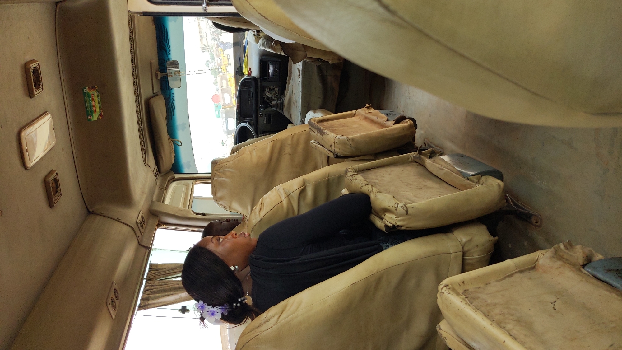 The image displays the interior of a passenger vehicle, likely a bus or large van, seen from a slightly elevated, rotated perspective. A woman with dark hair, adorned with small purple flowers and wearing earrings, a necklace, and a black long-sleeved top, is seated in a light-colored, worn upholstered seat on the right side of the vehicle. She appears to be resting or observing her surroundings during a journey.
The vehicle's interior features multiple rows of similar light-beige or cream-colored seats, some showing signs of significant wear and tear, with cushions folded up in the foreground revealing metal frames. The ceiling and side panels are also light-colored, equipped with overhead light fixtures or vents. On an overhead panel near the front, a small green wrapper or packet is visible, bearing the text "FRUITFULNESS SWEET ORANGE."
Through the front windshield, the driver's dashboard and steering wheel are discernible. A rearview mirror is attached to the windshield, which has a blue-tinted strip at the top. The bright light streaming in indicates it is daytime. Outside, other vehicles and what appears to be an urban or semi-urban street scene are visible, consistent with transit in Ado, Nigeria. The overall scene suggests a routine travel experience.