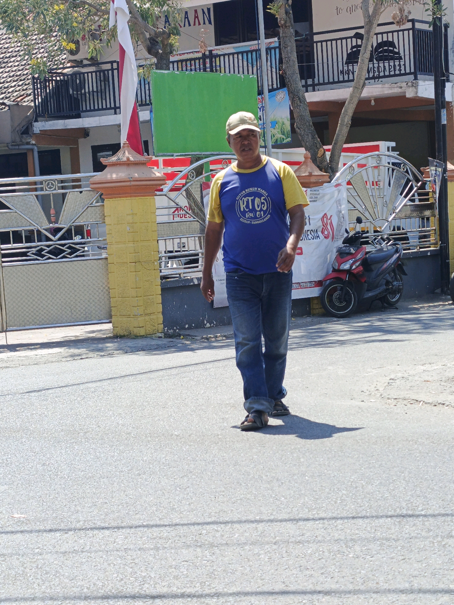 An adult male walks on a paved road in Ketegan, Indonesia. He is wearing a beige baseball cap, a blue t-shirt with yellow sleeves, and blue denim jeans. The t-shirt has a white circular logo with text and a design. He is also wearing sandals. Behind him, a metal fence with a yellow pillar and decorative elements separates the road from a building. The building has multiple stories, with balconies and windows visible. A red motorcycle is parked near the fence. A national flag, white and red, is visible on a flagpole attached to the fence. A green rectangular sign is also mounted on a pole near the fence. The sky is bright, suggesting daytime.