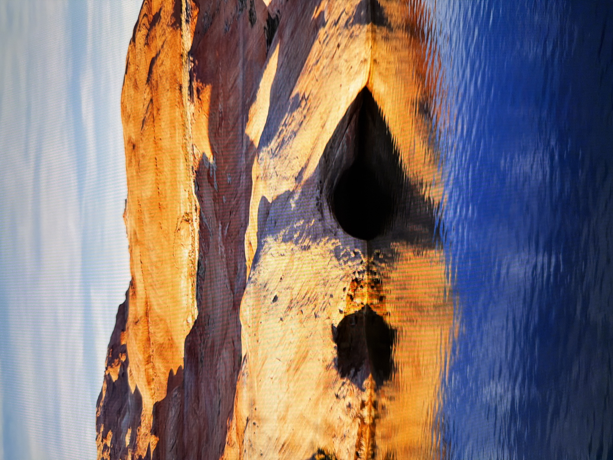 This is a landscape photograph of a desert canyon with a body of water in the foreground. The rocks are a warm orange and tan color, sculpted by erosion into smooth curves and sharp ridges. A dark cave-like opening is visible in the rock face at water level. The water is a deep blue and reflects the colors of the sky and the canyon walls, creating shimmering patterns. The sky above is a pale blue with thin, wispy white clouds. There are no people or any indication of urban environment. This image does not depict any location in Kyiv, Ukraine.