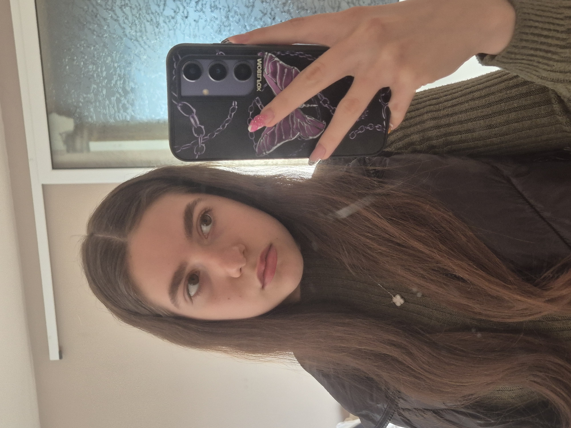 A young woman is taking a selfie indoors. She is holding a smartphone with a black phone case decorated with a purple butterfly and chain design. Her hair is long and brown. She is wearing a dark jacket and a green ribbed sweater. Light is coming from a window behind her, the glass has condensation or ice on it. The location is not discernible in this image. The location in the city of Pavlikeni, Bulgaria, cannot be determined from the provided image.