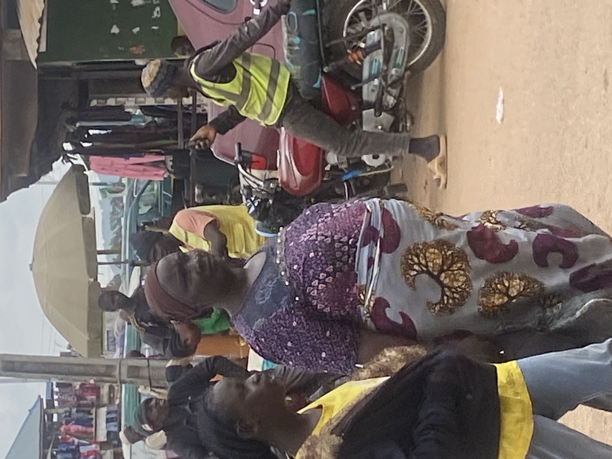 The image depicts an outdoor market scene in Bukuru, Nigeria. In the foreground, a woman with dark skin and a headscarf, wearing a purple patterned dress and a grey skirt with a large circular pattern, is looking to her left. To her left, a younger person with dark skin, wearing a yellow shirt and a black jacket, is also looking to their left. In the middle ground, a man wearing a yellow safety vest over a grey long-sleeved shirt and dark pants is seated on a red motorcycle, holding a mobile phone in his right hand. Behind him, other people and market stalls are visible, including clothing displayed on racks and an umbrella. The ground is dirt or gravel, and there is a pink car or vehicle on the right. The overall atmosphere suggests a busy marketplace.