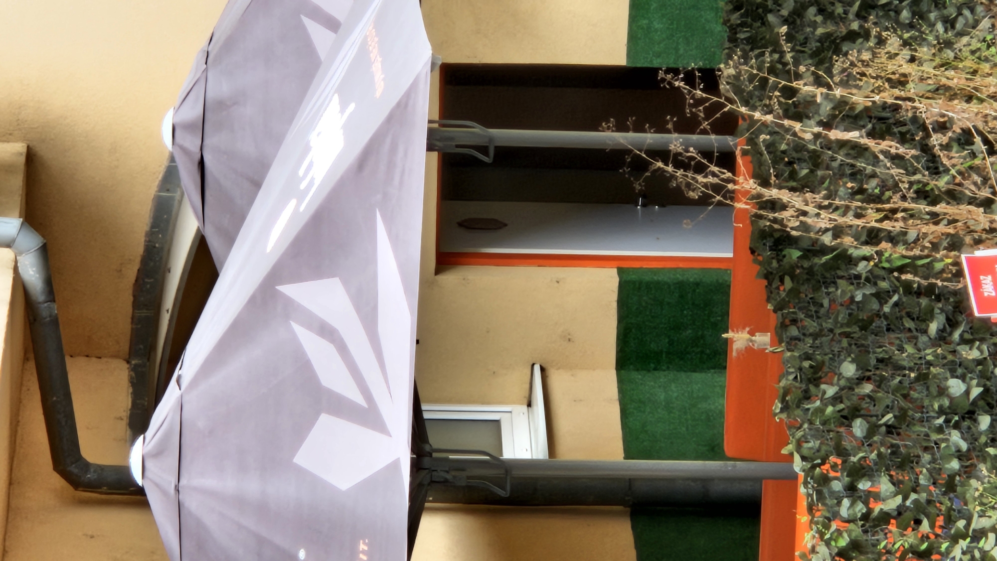 The image shows an outdoor seating area with two large, dark-colored umbrellas providing shade. The umbrellas have a white, geometric pattern. In the background, there is a building with a yellowish-beige facade, accented with dark green and orange sections. A downspout runs along the upper left side of the building. A window with a white frame is visible on the left side of the building, above a green-painted section. To the right of the window, an open doorway with an orange frame leads into a dark interior. In the foreground, there is an orange bench or railing, and a dense hedge of green foliage. To the right of the hedge, dry, dead plant stalks are prominent. A small red sign with white text, partially obscured by the foliage, is visible in the lower right corner, displaying the word "ZÁKAZ" which translates to "PROHIBITION" in Slovak. The overall scene suggests a cafe or restaurant exterior.