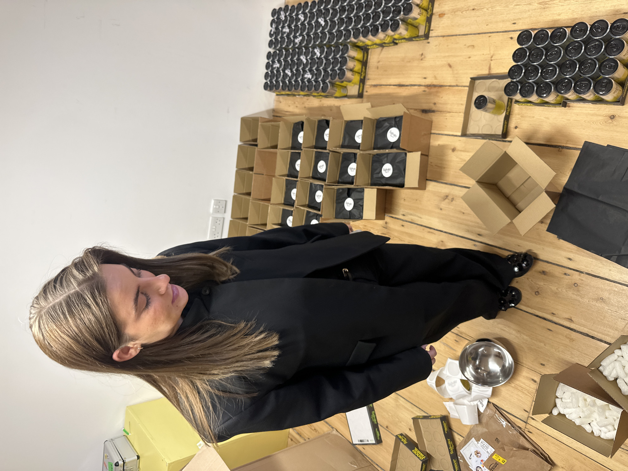 A high-angle photograph captures a female individual standing in an indoor commercial or storage space, identified as being in London, United Kingdom, by visible UK-standard electrical outlets on a white wall. The subject, possessing long brown hair, is dressed in a black suit jacket, black trousers, and black embellished flat shoes, positioned towards the left of the frame. She is looking downwards towards a collection of objects arranged on light wooden floorboards. The floor exhibits numerous open cardboard boxes, many containing individual items wrapped in black tissue paper sealed with white circular stickers. To the right, multiple arrays of yellow beverage cans with black tops are systematically organized in trays, bearing partial "Radwater" labeling. Other items present include opened cardboard boxes filled with white packing peanuts, a silver metallic bowl, rolls of white adhesive labels (some also marked "Radwater"), and brown paper envelopes, one displaying an "Amazon" logo. The scene indicates an active workspace dedicated to product packaging or distribution.