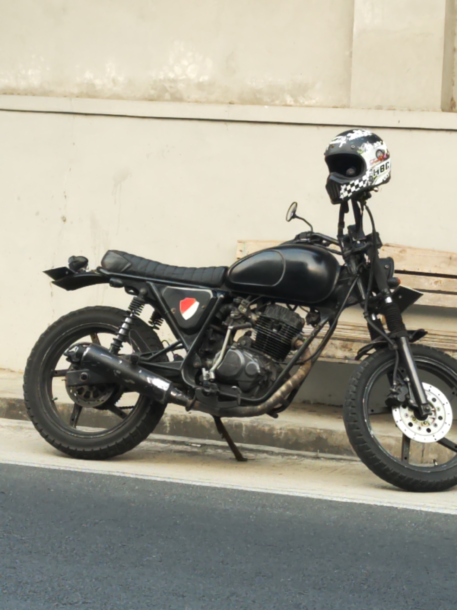 A black motorcycle with a black seat and chrome accents is parked on a sidewalk in front of a light-colored building. A black helmet with a white checkered pattern and various stickers is placed on top of the handlebars. The motorcycle has a black exhaust pipe and a red and white emblem on its side. The background consists of a textured wall and a wooden bench. The scene appears to be in Jaten, Indonesia, as the image is tagged with that location.