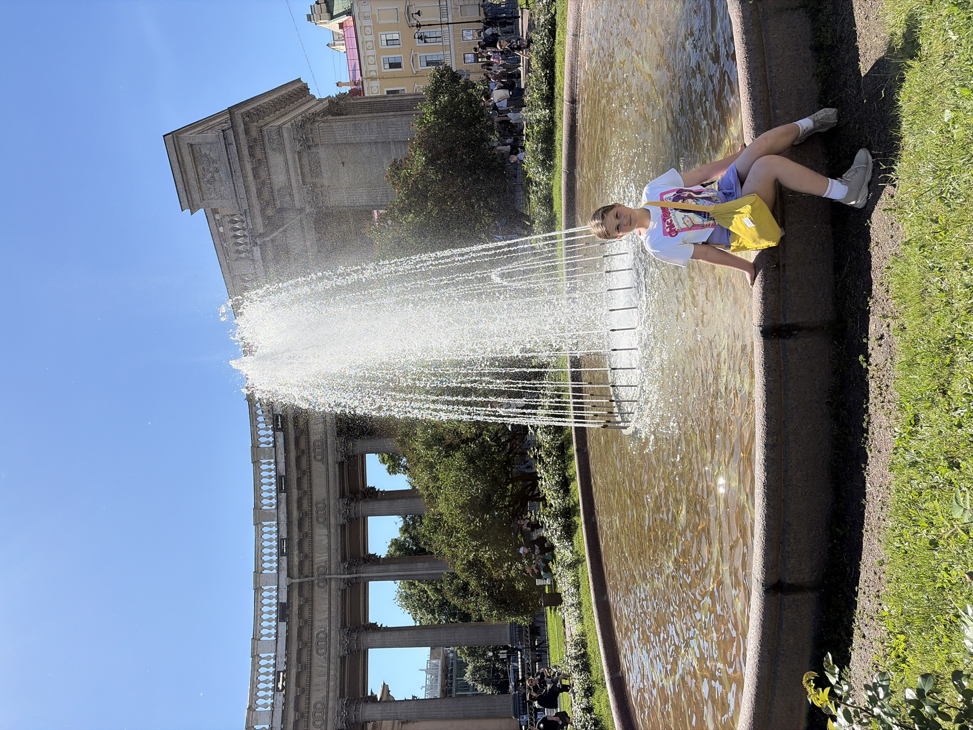 A young girl sits on the edge of a fountain, looking towards the camera and smiling. She is wearing a white t-shirt with a graphic print, purple shorts, white socks, and grey sneakers. A bright yellow bag is placed beside her. The fountain is large and ornate, with water spraying upwards. In the background, there is a grand, classical-style building with columns and intricate architectural details. Lush green trees and grass surround the fountain and building, suggesting a park or public square. Other people are visible in the background, some sitting on benches and others walking. The sky is a clear, bright blue, indicating a sunny day. The light suggests it is midday. The architectural style and the overall atmosphere point to a location in Saint Petersburg, Russia.