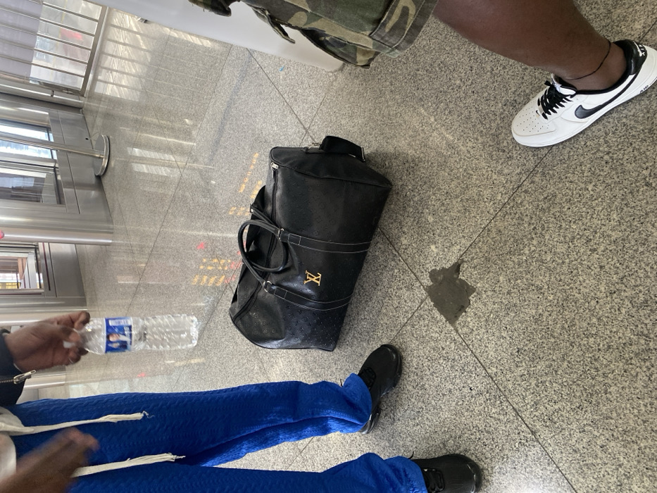 The image depicts an indoor scene with a speckled gray granite-ttiled floor. The setting is identified as the city of Zhongshan, China.
Three individuals are partially visible. On the left, a person is shown from the waist down, wearing textured bright blue pants with a white drawstring and black athletic shoes featuring a visible air sole. Their right arm and hand are visible, holding a clear plastic water bottle with a blue label displaying a portrait of a person.
A black duffel bag, bearing a gold-colored "LV" logo and an embossed pattern, rests on the floor between the individuals. Adjacent to the duffel bag and near the left individual's feet, a dark, irregularly shaped liquid spill is present on the tiles.
On the right side of the frame, a second person's lower leg and foot are visible, wearing camouflage-patterned shorts and white Nike Air Force 1 sneakers with black laces and black swooshes. A third individual is indicated by reflections in the background but is not directly visible.
In the background, shiny metallic structures, consistent with turnstiles or security gates, are visible. Reflections of these structures and an indistinct grid-like pattern are present on the polished floor and other surfaces, suggesting artificial or natural overhead lighting.