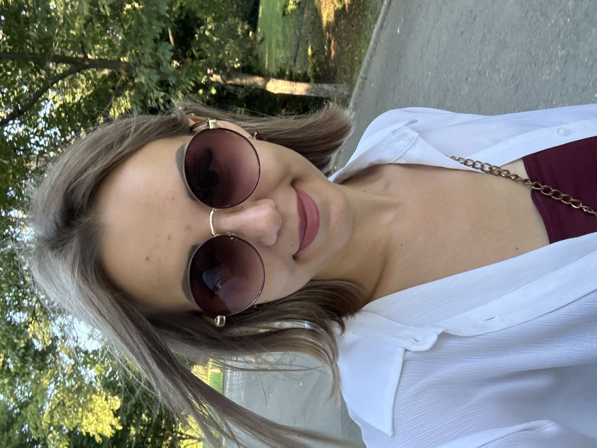 A young woman with light brown hair and fair skin, wearing large, round, gradient sunglasses and a white collared shirt, is photographed outdoors. Her hair is styled in a bob and appears windswept. She is smiling faintly and wearing a dark red top underneath her shirt. A gold chain accessory is visible over her shoulder. The background consists of trees with green foliage, suggesting a park or a natural setting within a city. The ground is paved, indicating a pathway or road. The lighting suggests it is daytime, likely late afternoon due to the warm tones and shadows. The specific location within Yuzhno-Sakhalinsk, Russia, cannot be determined from the visual information alone.