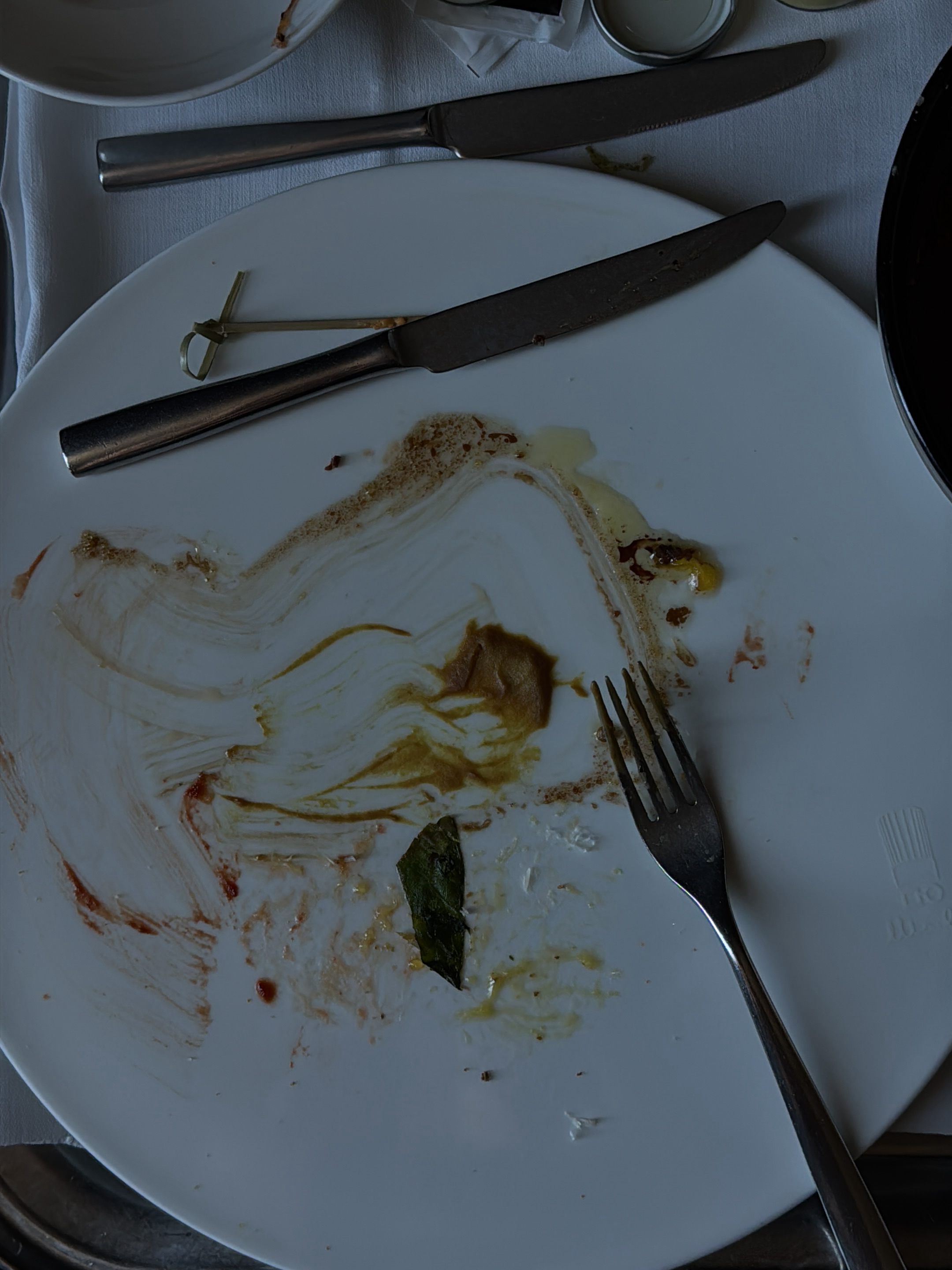 This is a top-down, close-up, dimly lit shot of a used white ceramic plate. The plate is stained with remnants of food, including brown sauces, a few dark red smears, and some yellow liquid. A single dark green leaf, possibly a herb, lies on the plate. Utensils are present: a metal fork with four tines is on the right side of the plate, and two metal knives are also on the plate. One knife is positioned horizontally across the top portion of the plate, and a small wooden skewer with a green tie is near its handle. The second knife is positioned diagonally in the lower half of the plate. The background is a white textured tablecloth, with glimpses of other tableware, including what appears to be a silver lid and a dark round object on the upper right. The scene does not provide any discernible indicators of a specific location in Milan, Italy.