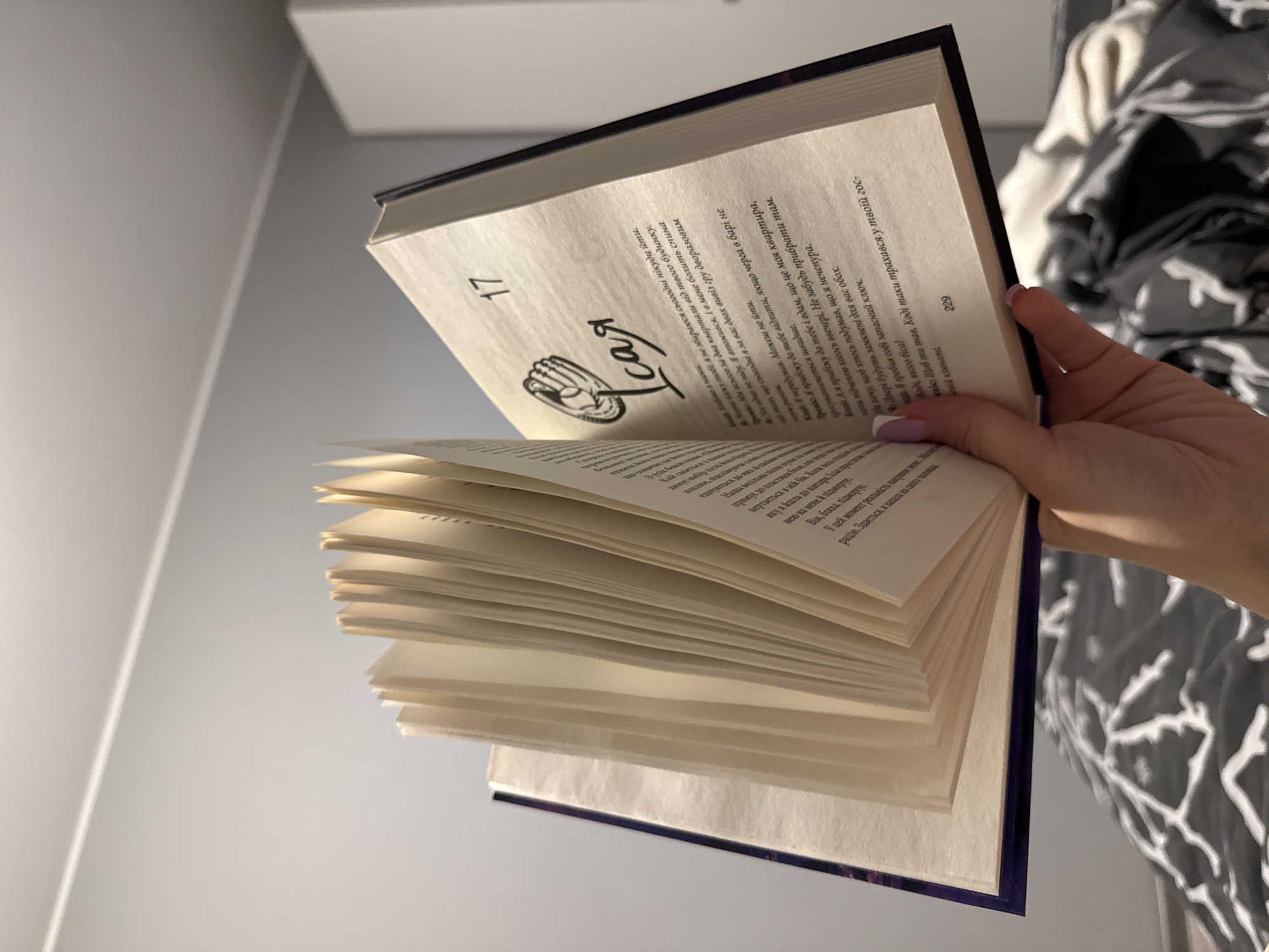 This is a close-up, indoor shot of an open book being held by a person's hand. The book is primarily white with purple binding visible. The pages are thick and cream-colored, with black text. The left page has the number "17" printed prominently near the top, followed by some text and a drawing of what appears to be a hand. The right page has the number "229" printed vertically along the right edge.
The person's hand, with neatly manicured nails painted light purple, is gently holding the right side of the book, its fingers splayed and resting on the pages. The background is softly blurred, showing a light gray wall on the left and a black and white patterned fabric, possibly a bedspread or blanket, on the right. The lighting suggests an indoor setting, possibly during the day with natural light contributing.
The visible text on the pages appears to be in Cyrillic script. The context provided states the location is Łódź, Poland, which is consistent with the use of Cyrillic. The book seems to be a novel or story, with chapter headings like "Ісая" (Isaiah) and page numbers. The content of the text suggests a narrative dialogue or internal monologue, hinting at a personal or fictional story being read. The overall mood is intimate and contemplative, focusing on the act of reading.