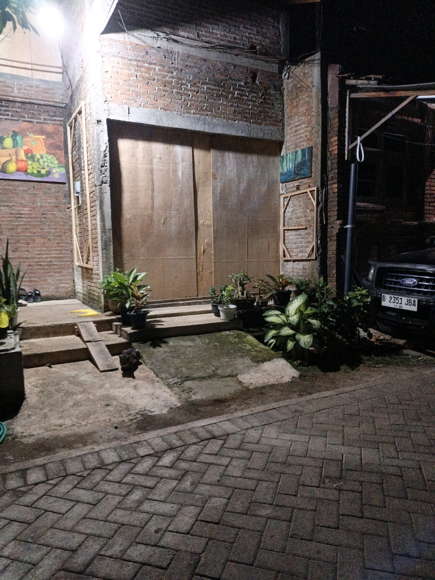 The image depicts an outdoor scene at night, possibly in the city of Cikal, Indonesia. In the foreground, a brick-paved walkway leads towards a building. To the right, the front of a dark-colored SUV with a license plate reading "B 2351 JBA" is visible. The SUV is parked next to a brick wall, with several potted plants situated in front of the wall and the vehicle.
Moving towards the building, there are concrete steps leading up to a large set of double doors made of what appears to be plywood. To the left of the steps, more potted plants are arranged, and a painting of fruit is hung on a brick wall. A small, dark-colored animal, possibly a cat, is seen near the base of the steps.
The building itself is constructed with exposed brick walls and concrete elements. Overhead, exposed wiring and lighting fixtures are visible. Another small painting, depicting a forest scene, is mounted on the brick wall to the right of the double doors. The overall impression is a somewhat rustic and utilitarian outdoor space, possibly a storage area or entrance to a residence or business.