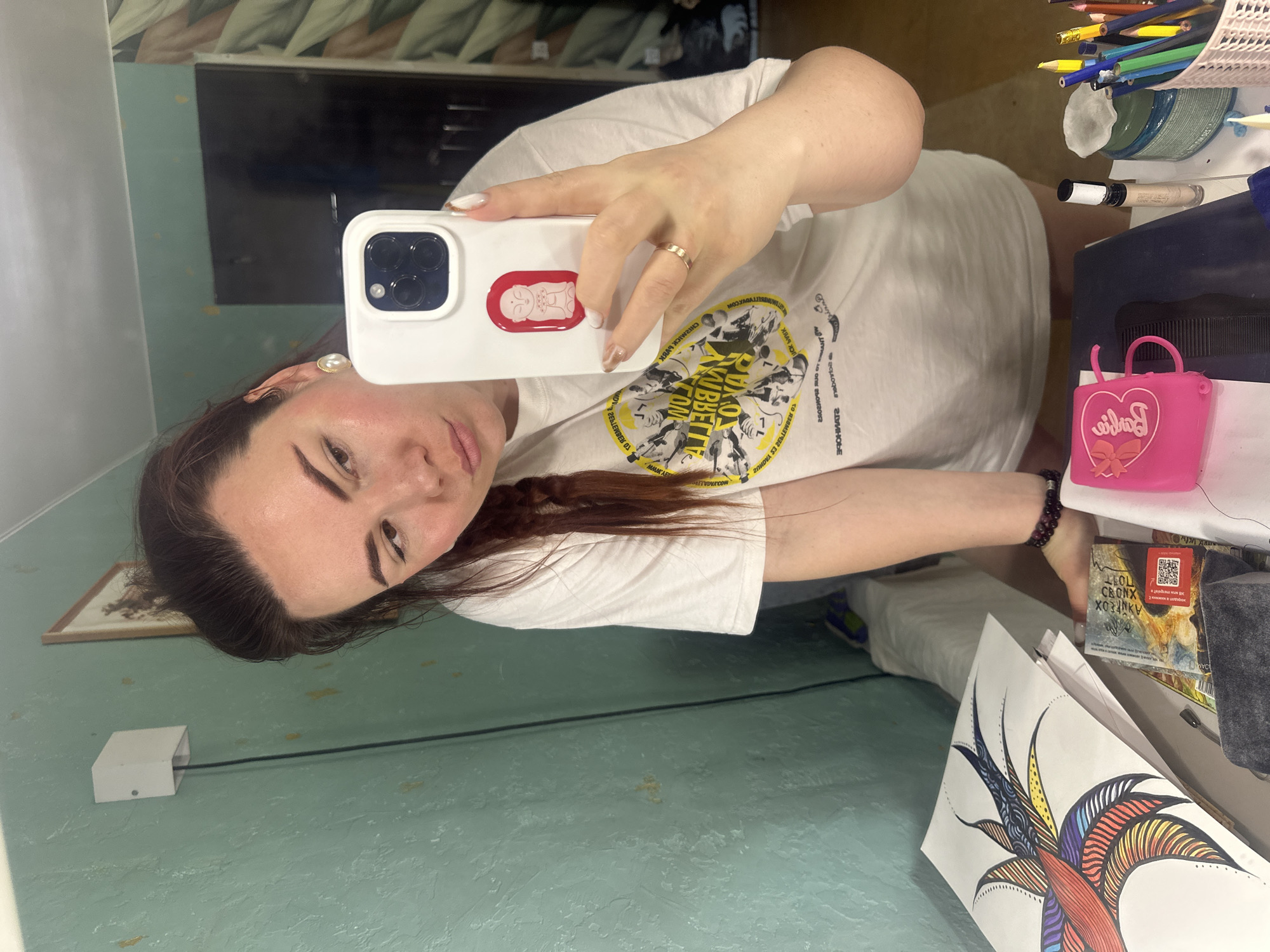 The image depicts a selfie taken by a woman with dark hair and fair skin. She is holding a white smartphone with a sticker of a baby in a red outline on the back. She is wearing a white T-shirt with a black and yellow graphic. A gold ring adorns the finger on her hand holding the phone.
The background includes a mirror reflecting a wall with a light fixture, and a desk cluttered with various items. On the desk, there are art supplies such as colored pencils in a container, a Barbie purse, a small grey box, and a drawing on paper with colorful line art.
Based on the OCR text "ЧЕРЁМУШКИ" visible on the box, the location may be in the Cheryomushki District of Moscow, Russia.