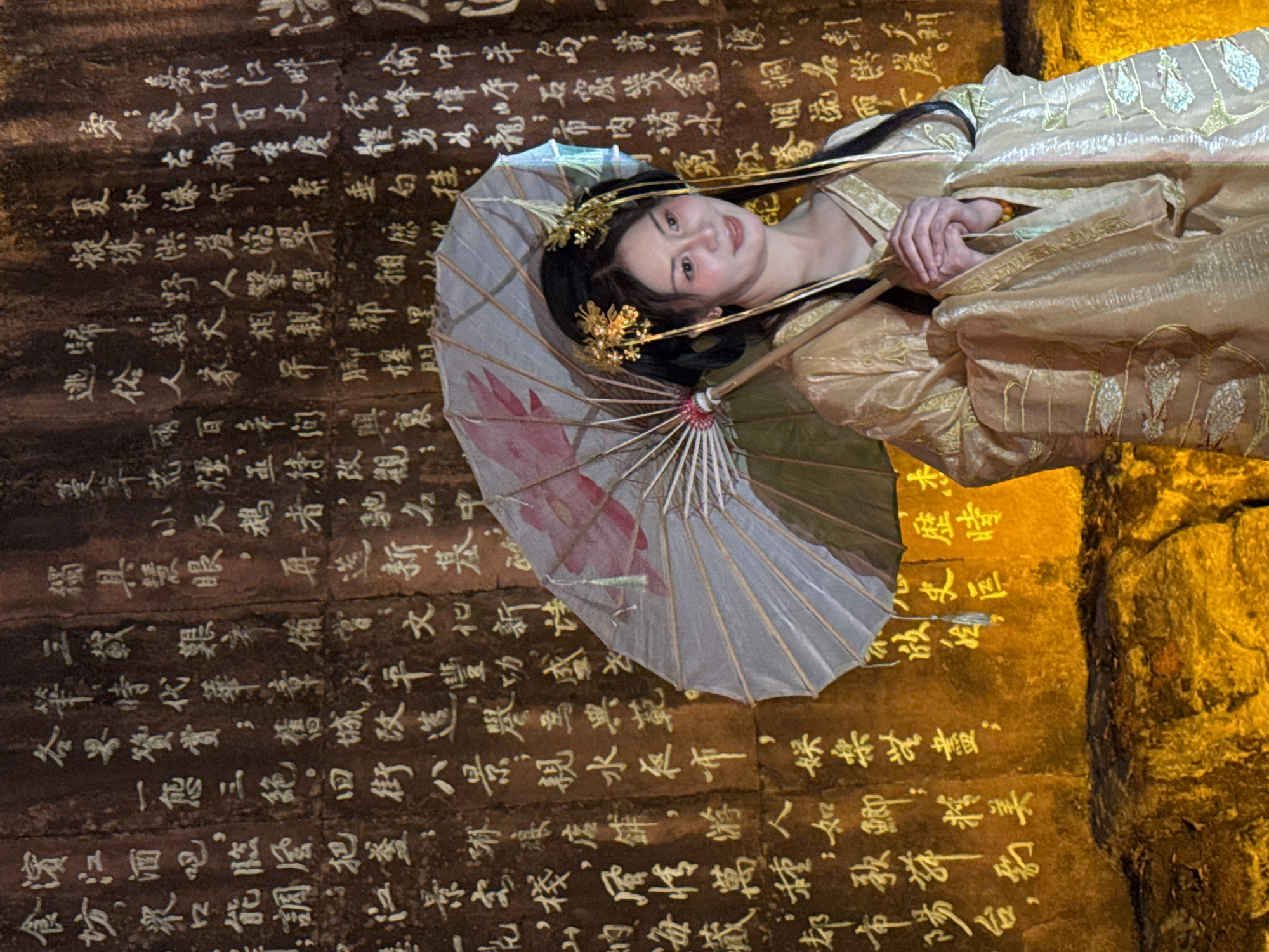 A woman in traditional Chinese attire stands in front of a stone wall carved with Chinese calligraphy, likely in Yuzhong District, China. The woman is holding an umbrella and her hands are clasped. The stone wall has ancient inscriptions in white against a dark background. The overall lighting suggests an evening or indoor setting.