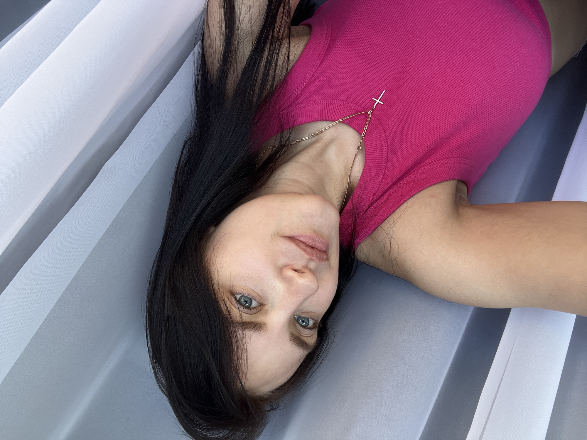 A woman with long, dark hair and blue eyes is captured in a selfie, wearing a bright pink ribbed tank top and a delicate gold cross necklace. Her head is tilted slightly to her left, and her gaze is directed towards the camera. She appears to be indoors, with a backdrop of vertically striped sheer curtains in varying shades of grey and white. The lighting suggests it is daytime. The specific location within Марковский, Russia, is not discernible from the visual information provided.