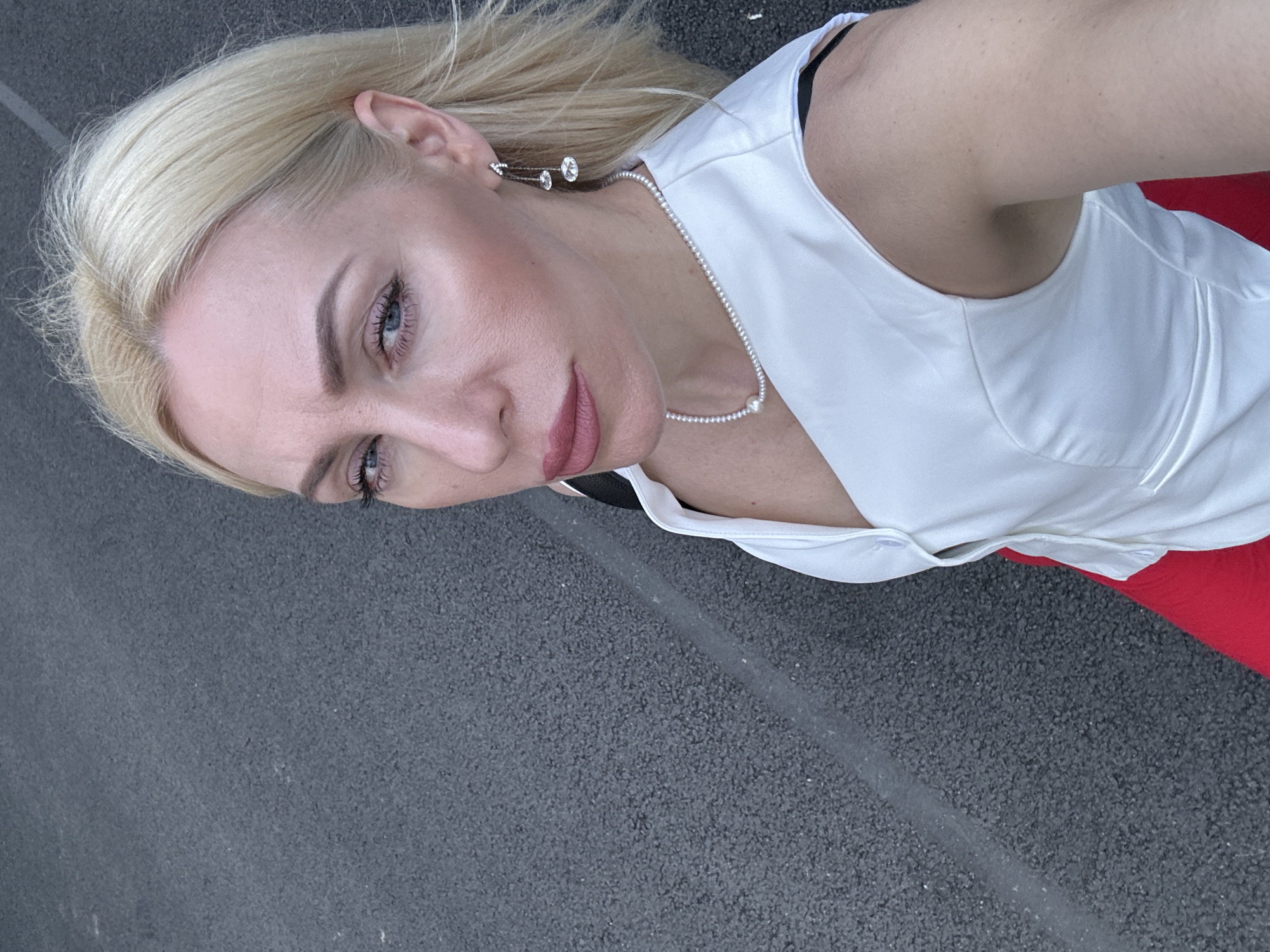 A blonde woman, with blue eyes, wearing a white sleeveless top, red skirt, a pearl necklace and dangling earrings, stands outdoors on a grey paved surface. The image is a selfie taken from a high angle, showing the woman from the chest up, with her face turned towards the camera. She has makeup on, including eyeshadow, eyeliner, and lipstick. The paved surface appears to be a road or a sports court, with a white line visible running diagonally across it. The background is out of focus, suggesting the photo was taken in an open area.