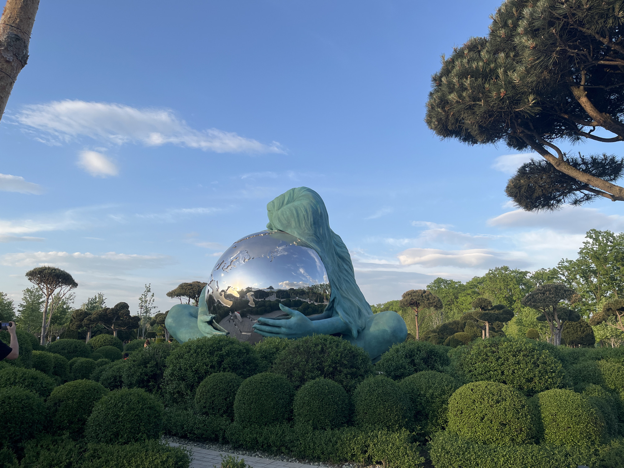A statue of a woman is sitting in a field in the city of Krasnodar, Russia. She is holding a large, silver sphere in her hands. The statue is surrounded by green bushes and trees. The sky is blue and cloudy.