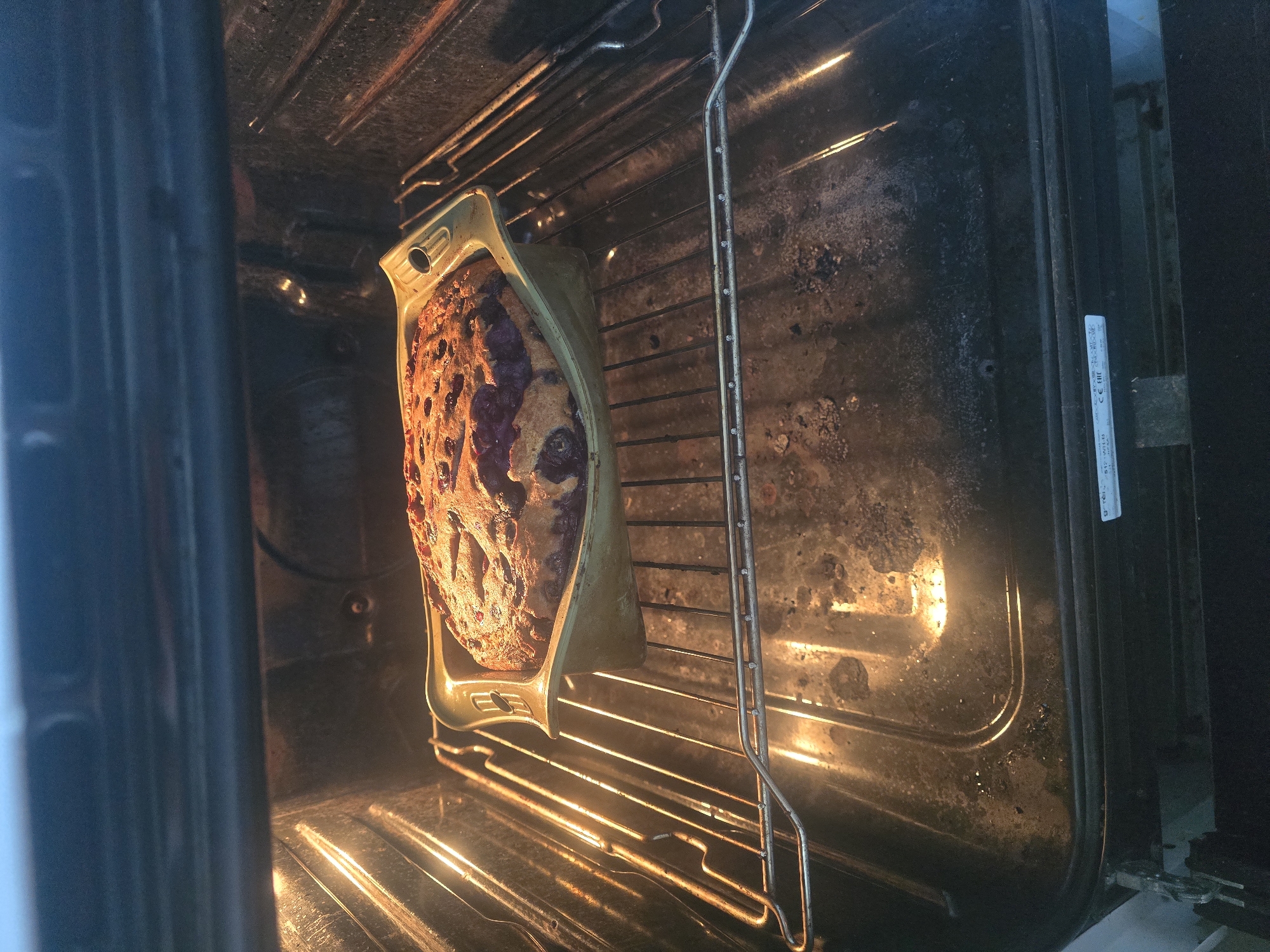 An overhead shot shows a loaf pan filled with a blueberry-studded baked good resting on the middle rack of a dirty oven. The oven light is on, illuminating the interior and the golden-brown crust of the baked good. The oven door is slightly ajar at the bottom, revealing a small portion of the oven's control panel and a label with the "CE" and "EAC" certification marks. The oven racks and interior surfaces are grimy, with visible burnt-on residue and stains.