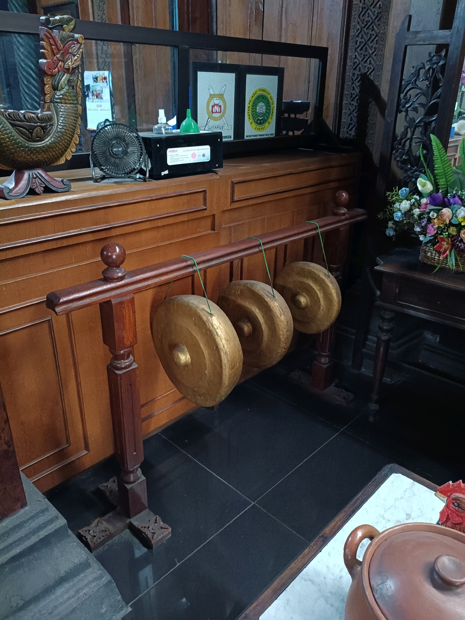 In the city of Purwomartani, Indonesia, a gong ensemble is displayed indoors. The ensemble consists of three bronze gongs suspended by green strings from a dark brown wooden rack. The gongs are positioned on a wooden floor with dark tiles. In the background, a wooden cabinet with intricate carvings is visible. On top of the cabinet, there is a small black fan, a spray bottle, a green funnel, and two framed certificates. To the right of the gongs, a dark wooden table holds a basket of colorful flowers. Further to the right, a portion of a traditional Indonesian house can be seen. A ceramic pot is placed on a marble-topped surface in the foreground. The scene appears to be in a cultural or historical setting.