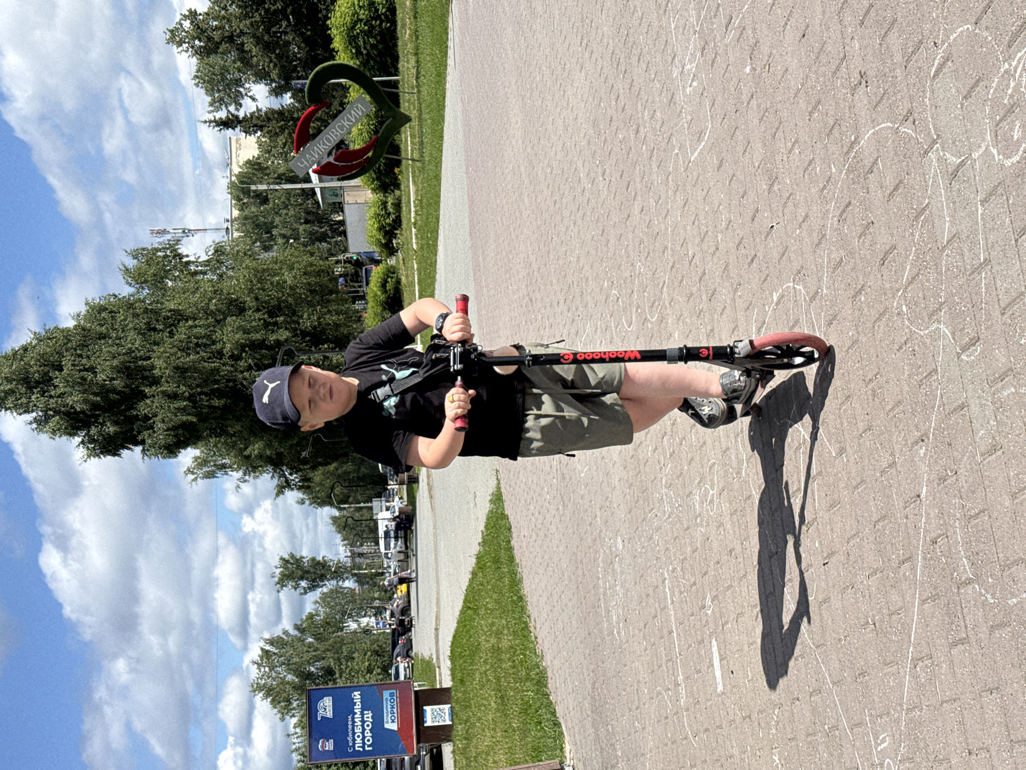 A young boy wearing a black t-shirt, olive green shorts, and a black baseball cap rides a red and black scooter on a paved walkway in Chaikovsky, Russia. The boy is facing forward, with his hands on the handlebars. His shadow is cast on the ground behind him. The walkway is made of brick pavers and has white chalk drawings on it, including a hopscotch grid. In the background, there are trees, buildings, and a sign with the text "С ЮБИЛЕЕМ, ЛЮБИМЫЙ ГОРОД!" (Happy Anniversary, Beloved City!) and "ВЛАДИМИР ЮРКОВ" (Vladimir Yurkov). Another sign shaped like a heart with text in Russian "ЧАЙКОВСКИЙ" (Chaikovsky) is visible in the background. The sky is blue with scattered clouds.