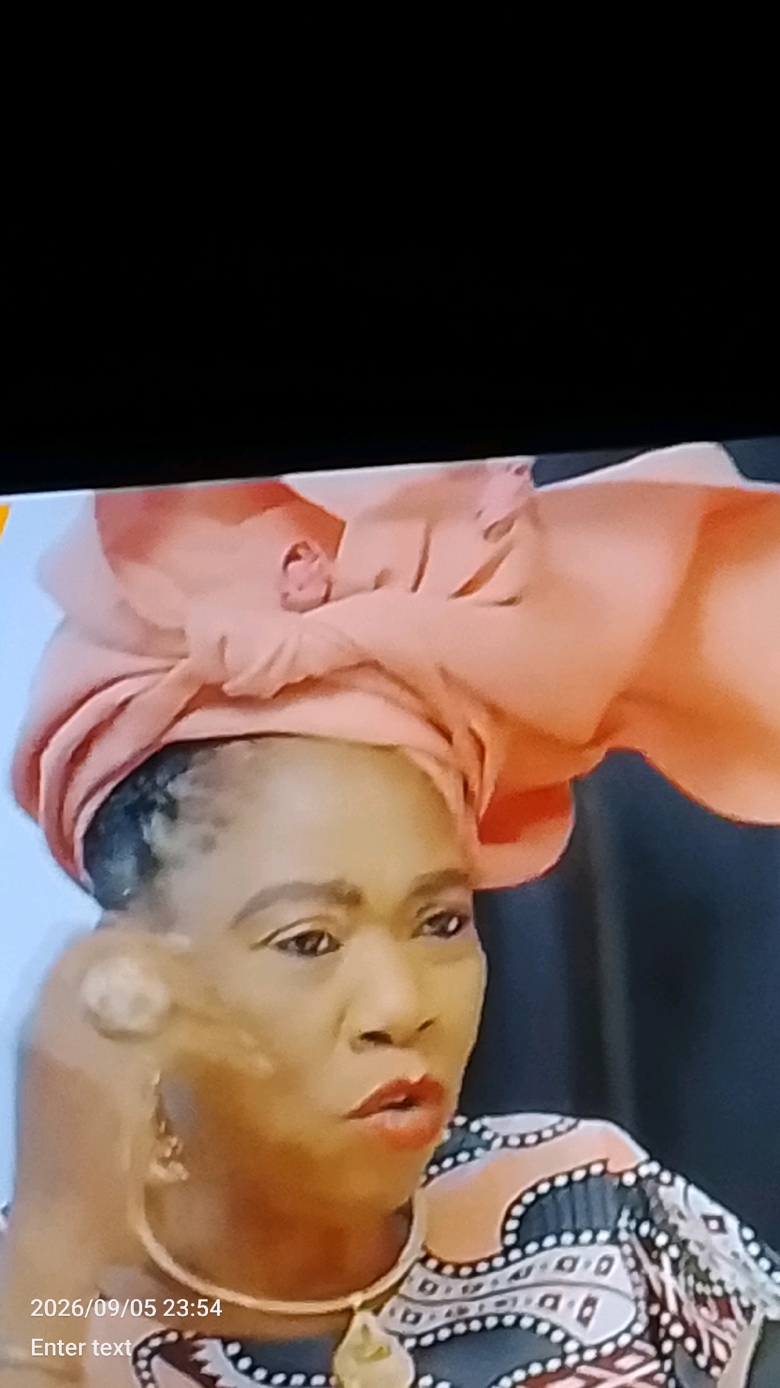 A medium shot captures a woman from Lokoja, Nigeria, from the chest up, looking to her right with her mouth slightly open. She is wearing a peach-colored headwrap, a pearl-like necklace, and a patterned top with geometric designs in black, white, and peach. Her left hand is visible near her face, and her right hand is not in frame. The background is dark and blurred, with a hint of a dark curtain or wall on the right. The timestamp "2026/09/05 23:54" and the text "Enter text" are superimposed on the lower left of the image.