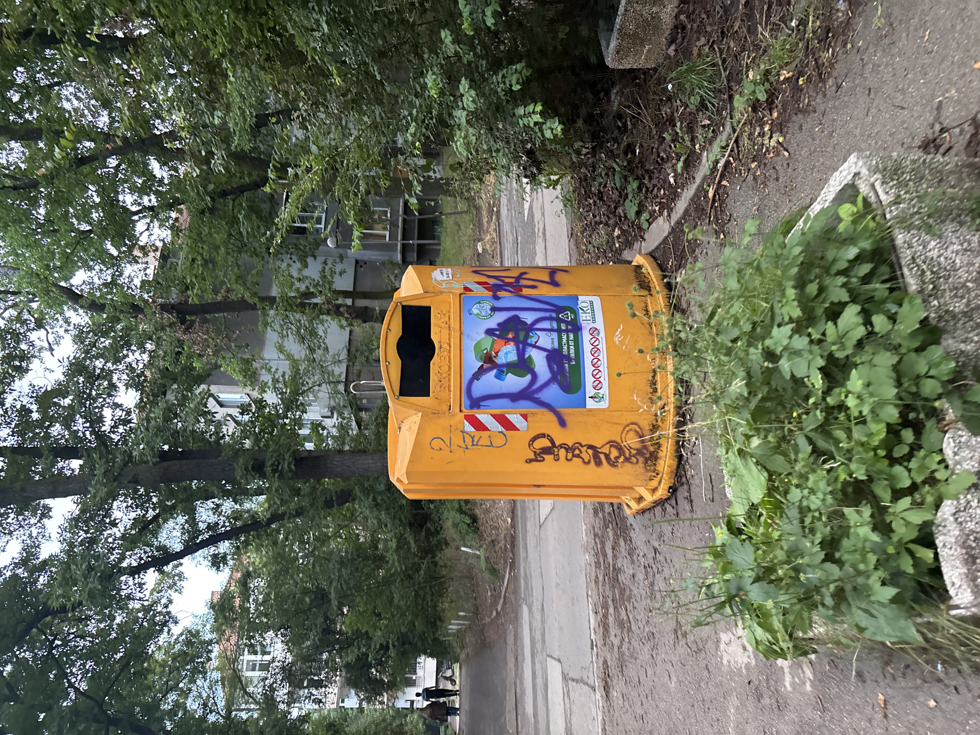 An eye-level shot shows a bright orange, cylindrical recycling bin situated on a paved pathway in a residential area of Sofia, Bulgaria. The bin is adorned with graffiti, featuring purple spray paint strokes over a cartoonish drawing and various symbols, including a red and white striped marker and a small logo with the text "EKO.BG". Text on the bin, partially obscured by graffiti, appears to be in Bulgarian and relates to recycling.
To the left of the bin, a paved pathway curves into the distance, bordered by trees and the facades of apartment buildings. Two individuals, one in a dark hooded jacket and the other in a dark shirt and jeans, walk away from the camera down the pathway, with a scooter visible between them. A third person, out of focus, is walking further down the path.
The environment is lush with vegetation. Green foliage and trees frame the scene, with dense bushes and weeds growing in front of the bin and along the edges of the pathway. The ground around the bin is covered with dry leaves and debris. The overall atmosphere suggests an outdoor, urban setting during daylight hours, possibly late afternoon given the overcast sky.