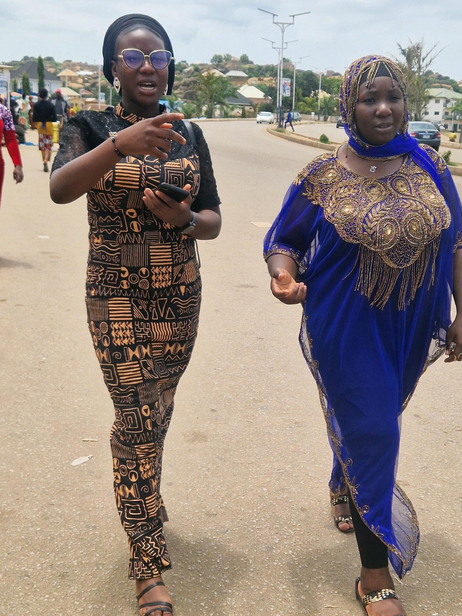 Two women are walking on a paved road in Kabong, Nigeria. The woman on the left is wearing a black headscarf, oversized sunglasses, and a black dress with an intricate tan geometric pattern. She is holding a smartphone in her left hand and pointing with her right hand. The woman on the right is wearing a blue hijab and a vibrant blue kaftan adorned with gold embroidery and beadwork. She is wearing black leggings underneath and has her right hand extended forward. The road appears to be a moderately busy street with buildings and some greenery visible in the background. The sky is overcast.