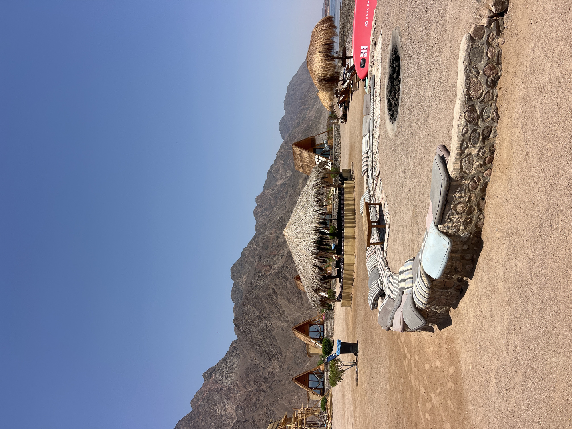 A sunny day unfolds in the resort area of Nuweiba, Egypt, showcasing a tranquil beachside scene. The foreground features a curved, stone-built seating area adorned with numerous cushions, some striped and others solid in varying colors. A wooden table is positioned in the center of this seating arrangement. Adjacent to it, a circular fire pit is visible, filled with dark stones.

Beyond the seating area, several structures with thatched roofs dot the sandy landscape. One prominent structure is a circular hut, likely a cafe or lounge, with seating arranged around it. To the left, three triangular, A-frame cabins with wooden exteriors and glass fronts stand out, suggesting accommodation units. A small, green tree and a blue trash can are positioned between these cabins and the larger hut.

In the middle ground, a raised platform with a thatched roof serves as a shaded seating area with cushioned benches. Further to the right, two sun loungers are placed under another thatched umbrella, near the water's edge. A bright red paddleboard, labeled "AQUA MA," rests on the sand. The background is dominated by rugged, brown mountains, characteristic of the desert landscape in this region. The clear blue sky stretches across the top of the image, indicating a cloudless day. The sea is visible on the far right, meeting the shore with a rocky edge. The overall impression is one of a peaceful, rustic beach resort.