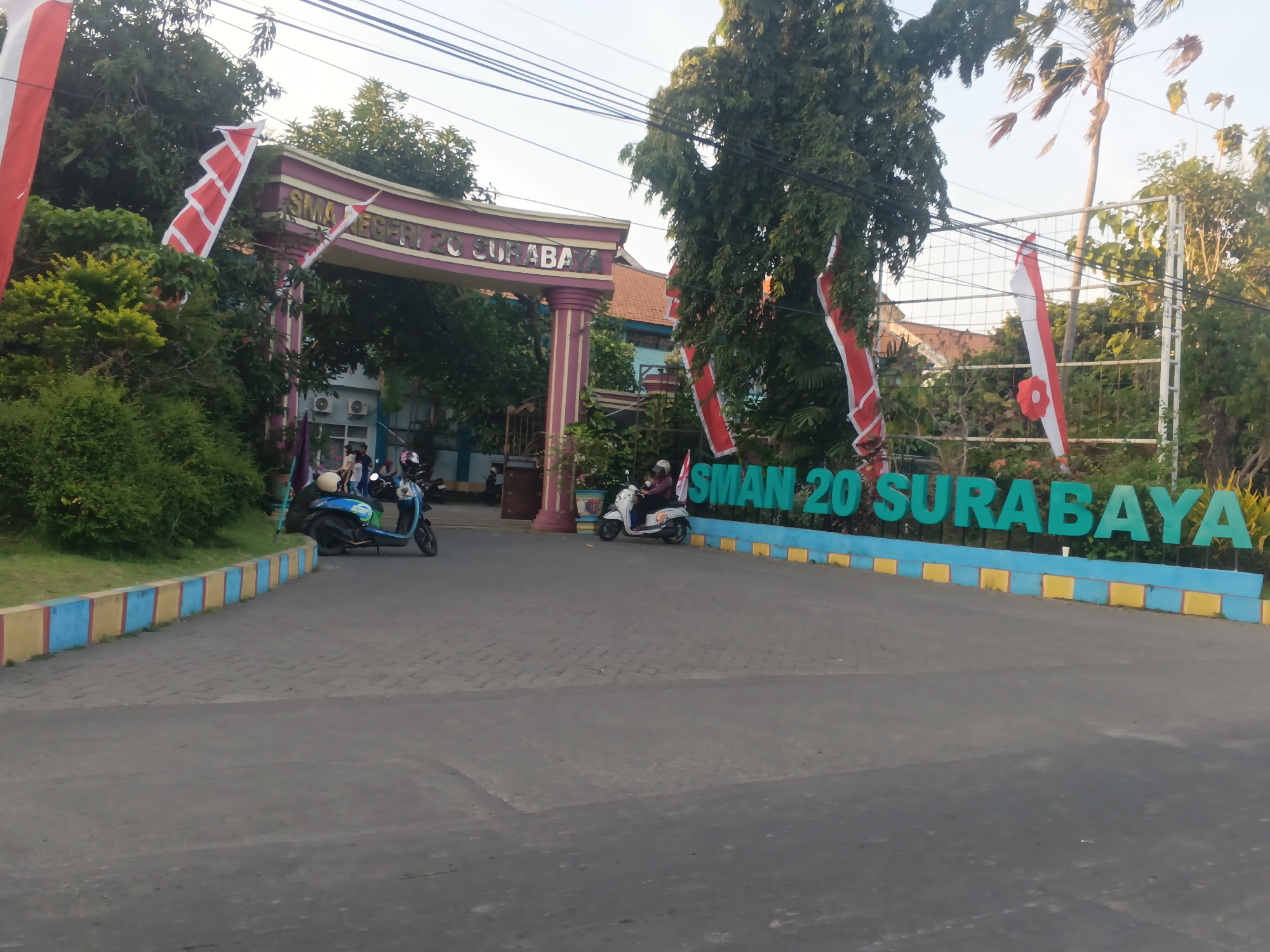 The image shows the entrance of "SMAN 20 SURABAYA," a high school in Surabaya, Indonesia. The school gate is adorned with Indonesian flags. In front of the gate, there are several motorcycles, two of which are being ridden by individuals. The school building is visible behind the gate, with trees and foliage surrounding the area. The ground is paved, and a decorative border of blue and yellow blocks lines the driveway. The sky is clear and appears to be late afternoon or early evening.