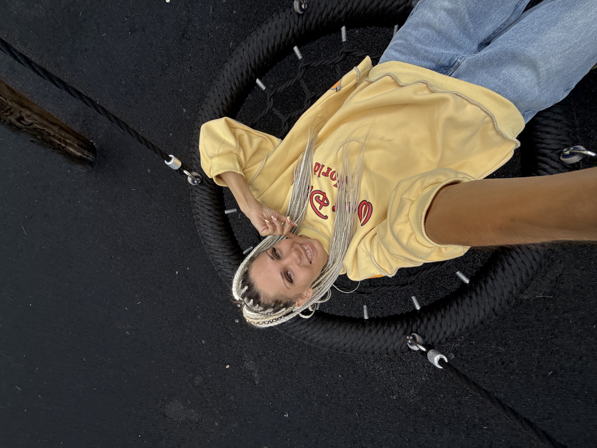 A woman with blonde braided hair is lying on a large, round, black rope swing. She is wearing a yellow sweatshirt and blue jeans. The swing is suspended by ropes attached to a dark surface, which appears to be a playground with a black rubberized surface. A wooden beam is visible in the upper right corner of the image. The woman is looking at the camera and smiling. The image is taken from a high angle, looking down at the woman and the swing. The location is identified as Rusanovo, Russia.
