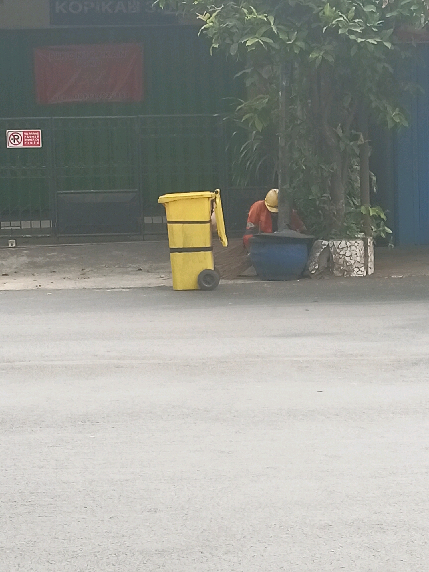 In this image, a person wearing an orange work jacket and a yellow hard hat is partially visible, crouching next to a large blue barrel. The scene appears to be an outdoor urban environment, likely on a sidewalk or street. To the left of the person, a bright yellow trash can with black stripes stands on wheels. Behind the trash can and the person, a dark green metal fence stretches across the background. Above the fence, a faded red banner with Indonesian text is visible. A small "No Parking" sign with a red circle and diagonal line is affixed to the fence. A large tree with lush green leaves is situated behind the blue barrel and the person, obscuring much of the background. The ground in front of the fence is a mixture of concrete and dirt. The foreground consists of a paved road or street, blurred due to motion. Based on the presence of Indonesian text on the banner and the overall aesthetic, the image is likely taken in Malang, Indonesia.