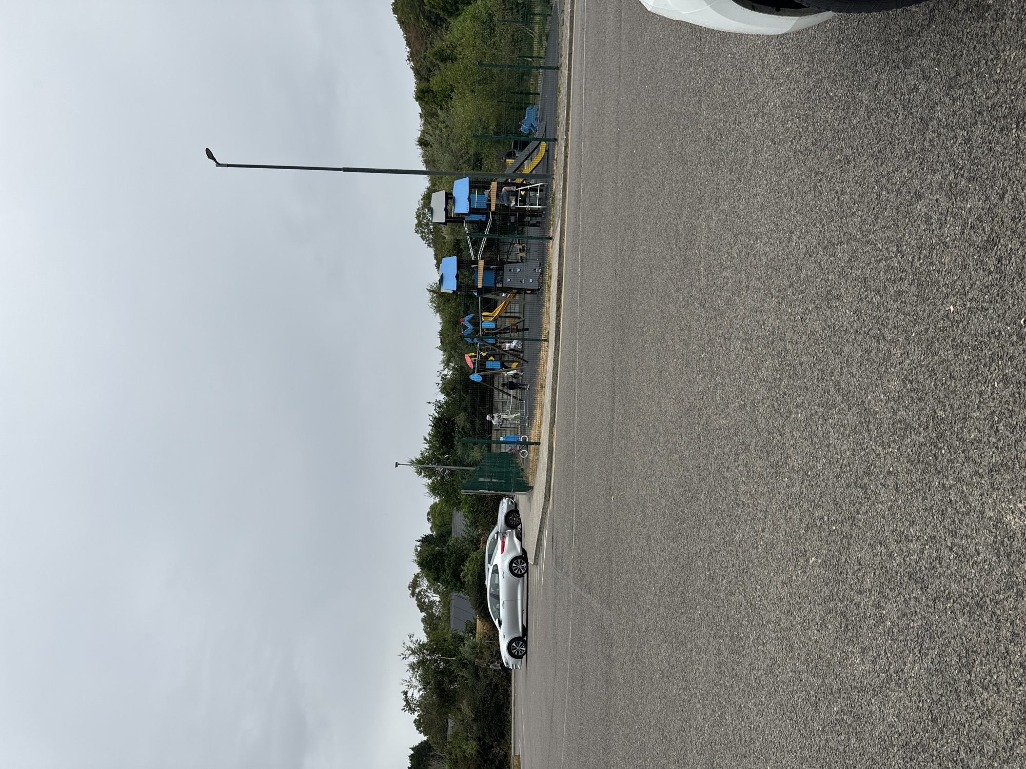 The image shows a playground enclosed by a green metal fence, located in Courtown, Ireland. The playground features blue and yellow climbing structures, slides, and a climbing wall. A few children and adults are visible within the fenced area. Outside the fence, to the left, are two silver cars parked in a paved area. A street lamp is visible in the background to the right of the playground. The sky is overcast, and the surrounding environment consists of trees and green foliage. The foreground is a grey asphalt surface, consistent with a road or parking lot.