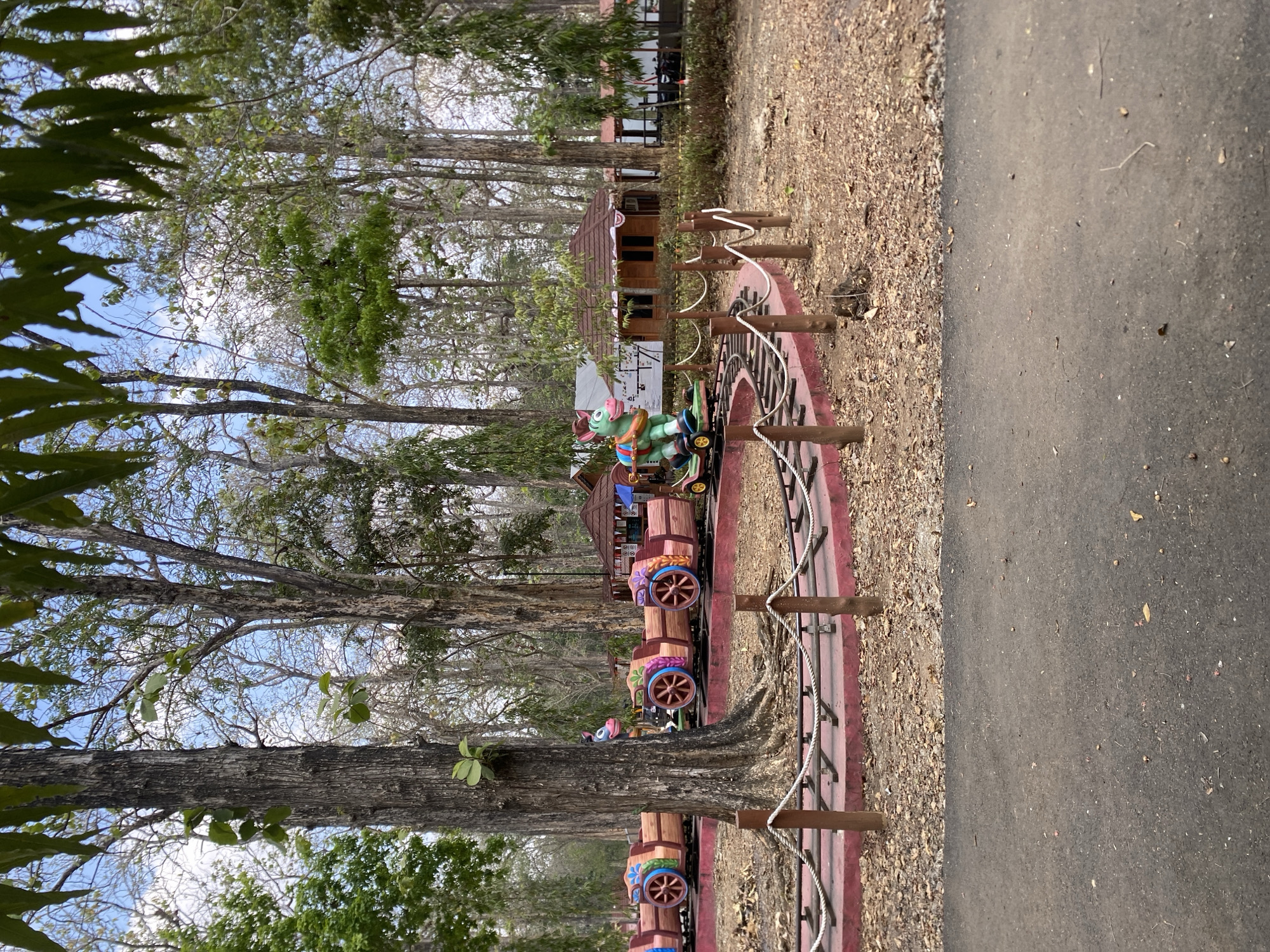 This image depicts a children's train ride in a park setting located in Ketileng, Indonesia. The train is composed of several colorful, individual cars, each with a wooden appearance and decorative wheels. One car features a green cartoon horse with a blue saddle, which is the lead vehicle of the train. The train is on a red-colored track that curves through a wooded area with many tall trees. The ground around the track is covered in dry leaves and dirt. In the background, there are small, brown wooden buildings with red roofs, resembling market stalls or small shops. The sky is visible through the trees, showing a mix of blue and white clouds. The foreground of the image shows a paved path or road. There are no people visible in the scene, suggesting the park is either closed or the ride is not in operation at the moment.