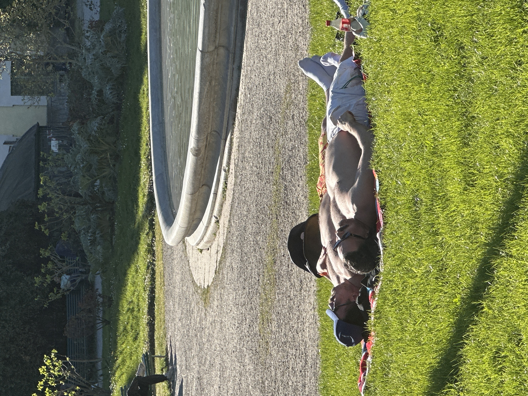 A man lies topless on a grassy area next to a fountain and a path in what appears to be a park setting. He is wearing light-colored pants, a dark baseball cap, and sunglasses. There is a blanket or other fabric beneath him. In the background are trees, buildings, and park benches. The scene suggests a sunny day in a public park. The image likely depicts a scene in Sempione Park in Milan, Italy, based on the visible architecture and park elements.