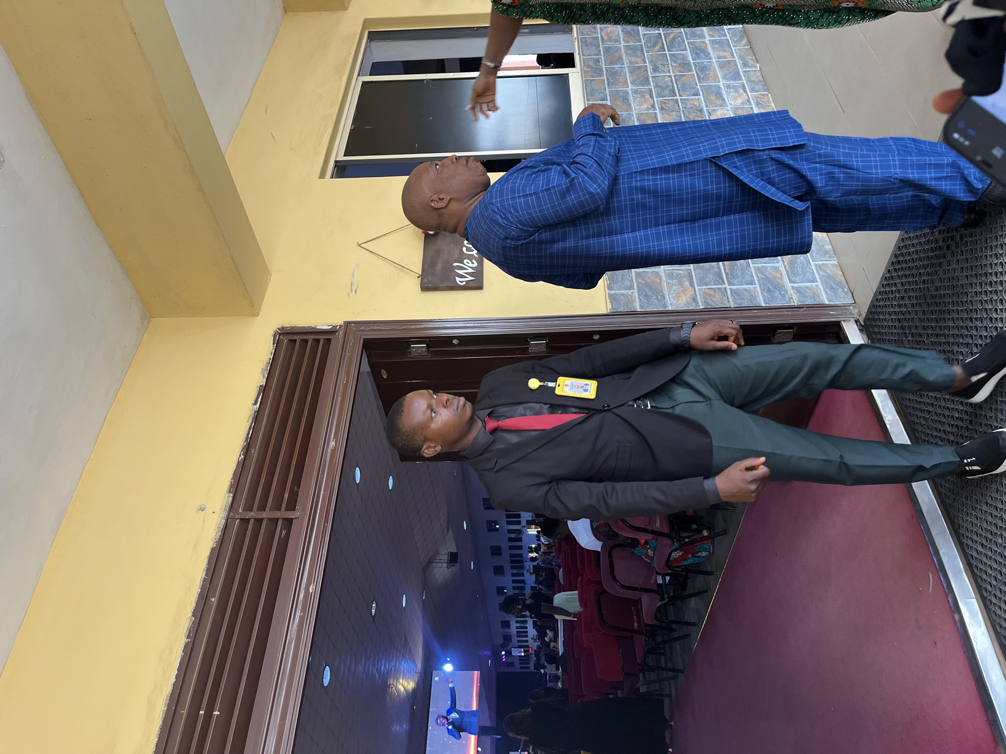 The image depicts two men in formal attire standing near the entrance of a building, likely in Abuja, Nigeria. One man is wearing a black suit with a red tie and a yellow ID badge. The other man is wearing a blue checkered suit. A woman's arm is visible above the man in the blue suit. Behind the man in the black suit, rows of red chairs can be seen inside the building, suggesting it may be a church or auditorium. A stage with a person presenting on a screen is visible at the front of the room. A welcome sign is on the building. The environment suggests a formal event or gathering is taking place.