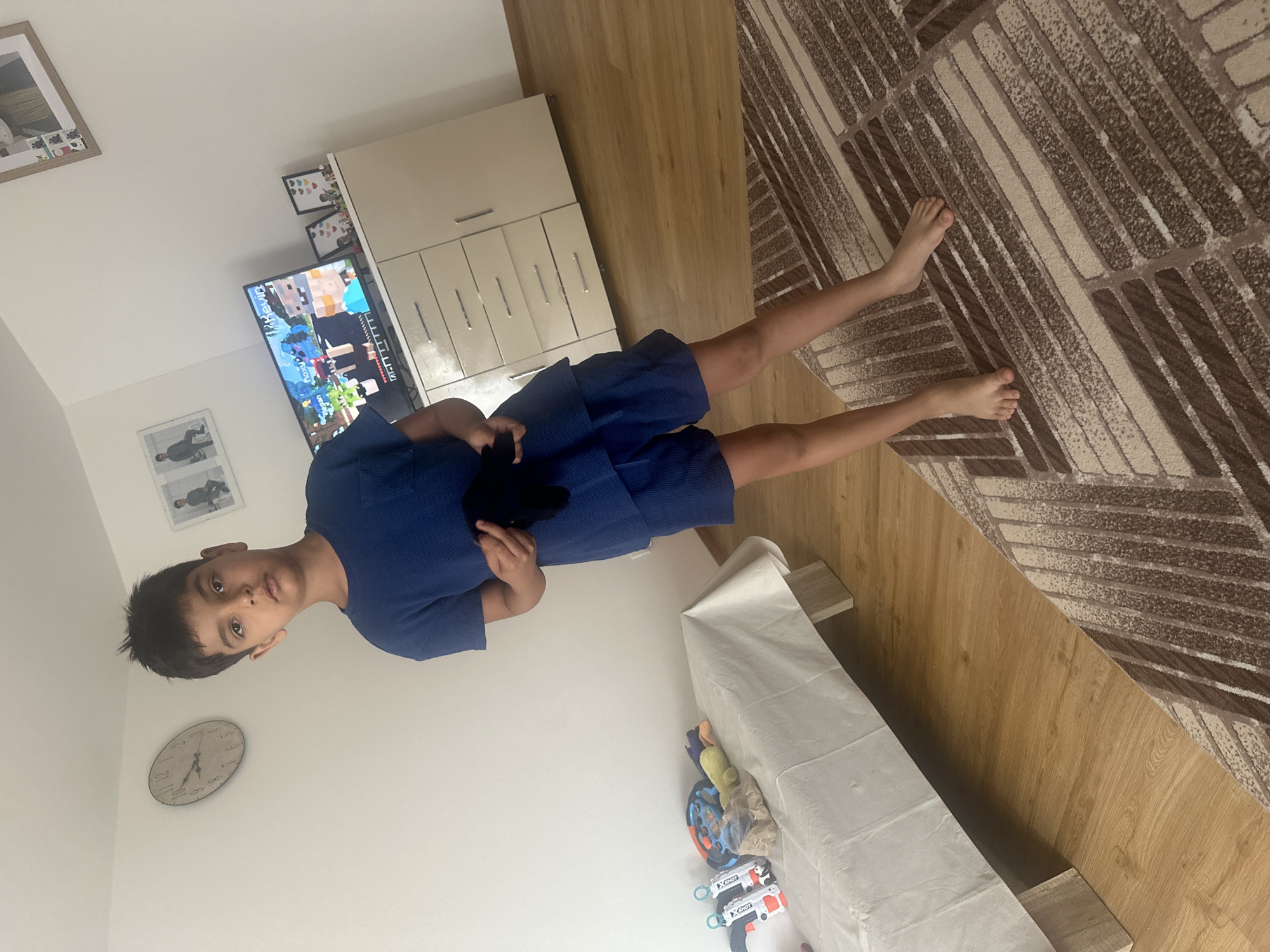 In an indoor setting, a young boy with dark hair and brown eyes, wearing a blue t-shirt and shorts, stands in the center of the frame. He holds a black object in his hands. The room features wooden flooring and a patterned rug. To the left, a bed is covered with a white cloth, and several toys are placed on it. A clock is mounted on the wall above the bed. To the right, a dresser with drawers and cabinets is visible, and a television screen displays a video game. Framed pictures are also present on the wall and on top of the dresser. The scene appears to be a child's bedroom or playroom. This image was captured in Mainburg, Germany.