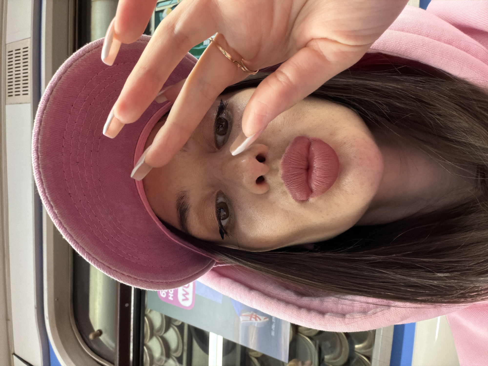 A young woman in a pink baseball cap and hooded sweatshirt poses in what appears to be a railway car, possibly the Saint Petersburg Metro, judging by the Cyrillic text on the signage behind her. She holds her hand up to her face, displaying long, manicured nails. A gold ring is visible on one of her fingers. Her hair is dark brown. The focus is on her face, particularly her lips, which are prominent. The background shows a partial view of the railway car interior with windows, seating, and signage. The overall lighting suggests it's daytime.