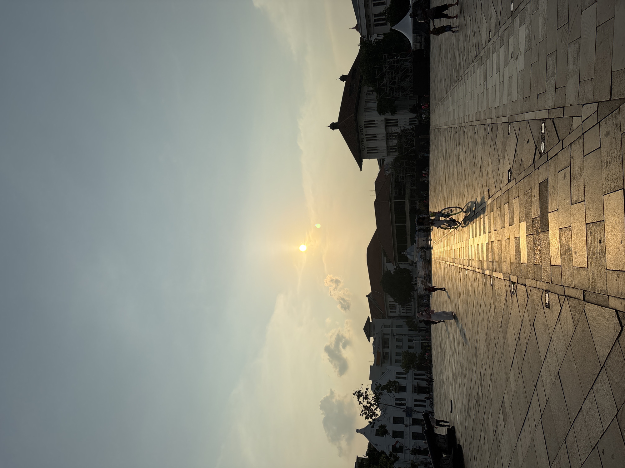 This image depicts Fatahillah Square in Kota Tua, West Jakarta, Indonesia, during a sunset. The sun is low in the sky, casting a warm, golden light across the square and creating long shadows. The square is paved with stone tiles, and a pathway illuminated by the setting sun runs through the center.
Several people are visible in the square. In the foreground, a person is riding a bicycle, their silhouette casting a long shadow on the ground. Further back, a person in a headscarf walks across the square, and another individual, possibly a child, walks beside them. In the distance, other figures are scattered throughout the plaza, engaged in various activities.
The background features several colonial-era buildings with distinctive architecture, including white facades and red-tiled roofs. Trees are interspersed among the buildings, and some scaffolding is visible, suggesting ongoing construction or maintenance. A large cannon, a historical artifact, is positioned on the left side of the frame. Tents are set up on the right side of the square, possibly for vendors or events. The sky is a pale blue with some scattered clouds illuminated by the setting sun.