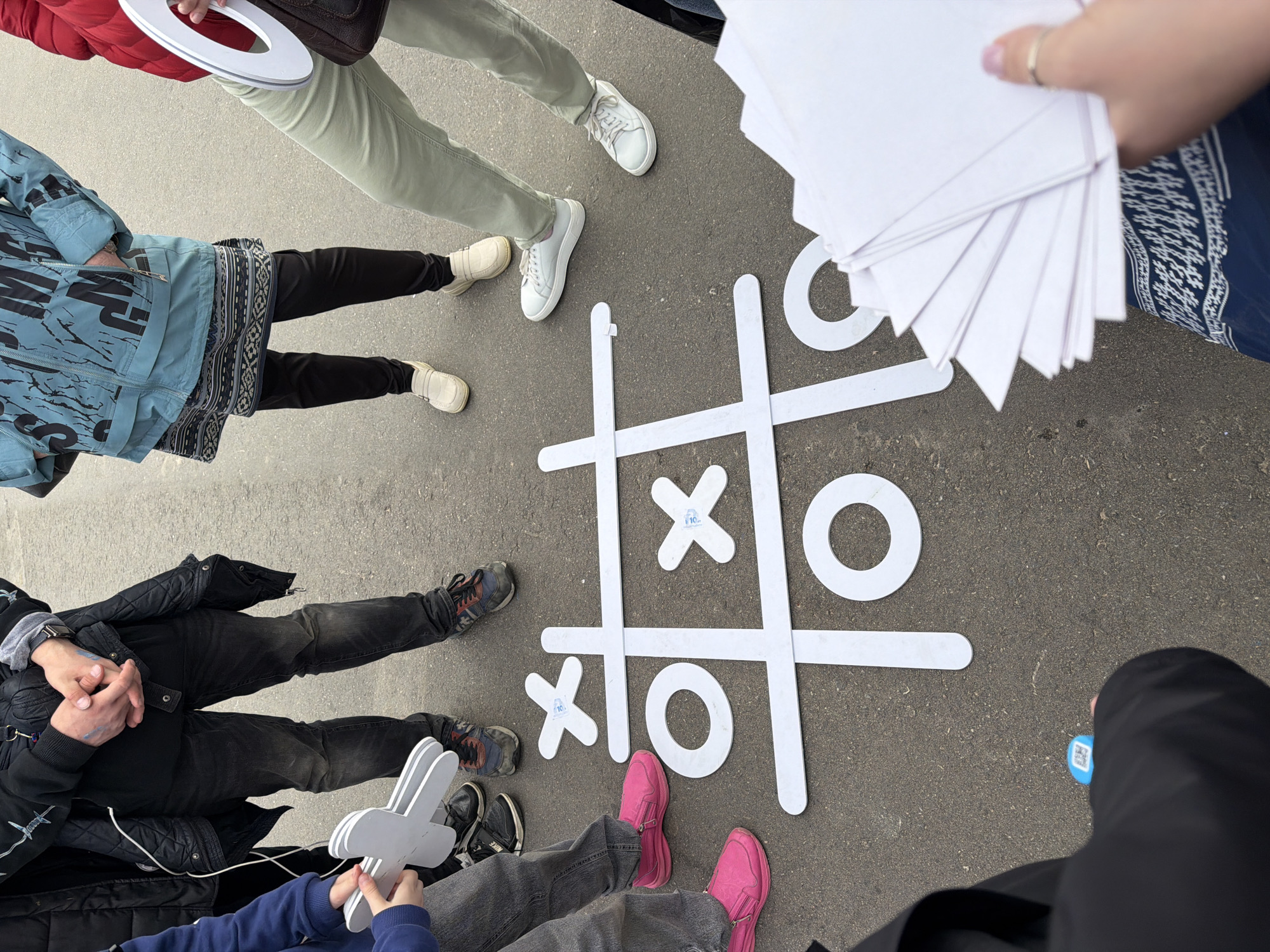 The image depicts a group of people standing around a large tic-tac-toe board made of white material on a paved surface. The board contains two "X"s and two "O"s. The people are dressed in casual clothing, with different styles and colors of pants, jackets, and shoes. One person is holding a stack of white papers. It is implied that this image was taken in the city of Moscow, Russia.