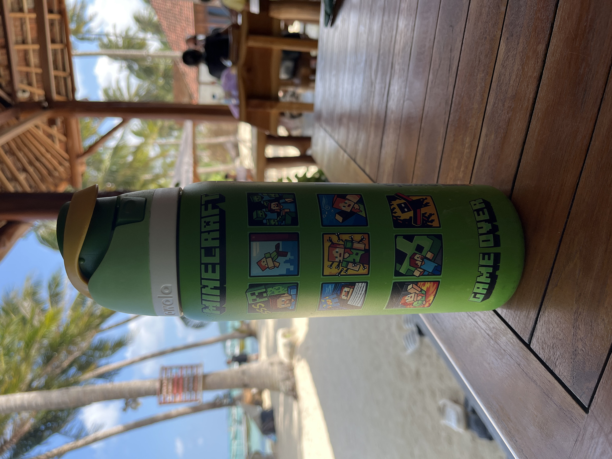 A green, insulated water bottle featuring Minecraft graphics rests on a wooden table. The bottle has a yellow lid and a white band below the lid. The Minecraft graphics depict characters and scenes from the game, arranged in a grid. Below the graphics, the text "GAME OVER" is printed in green block letters.
The bottle is situated outdoors at what appears to be a beachside cafe in Legonipah, Indonesia, as indicated by the background. In the background, palm trees sway under a bright blue sky with scattered clouds. Beyond the palm trees, a sandy beach meets the turquoise ocean. Several thatched-roof structures, typical of tropical resorts or restaurants, are visible. People are seated at tables in the background, some appearing to be patrons or staff. The wooden table the bottle sits on has a slatted surface. The overall atmosphere suggests a relaxed, tropical vacation setting.