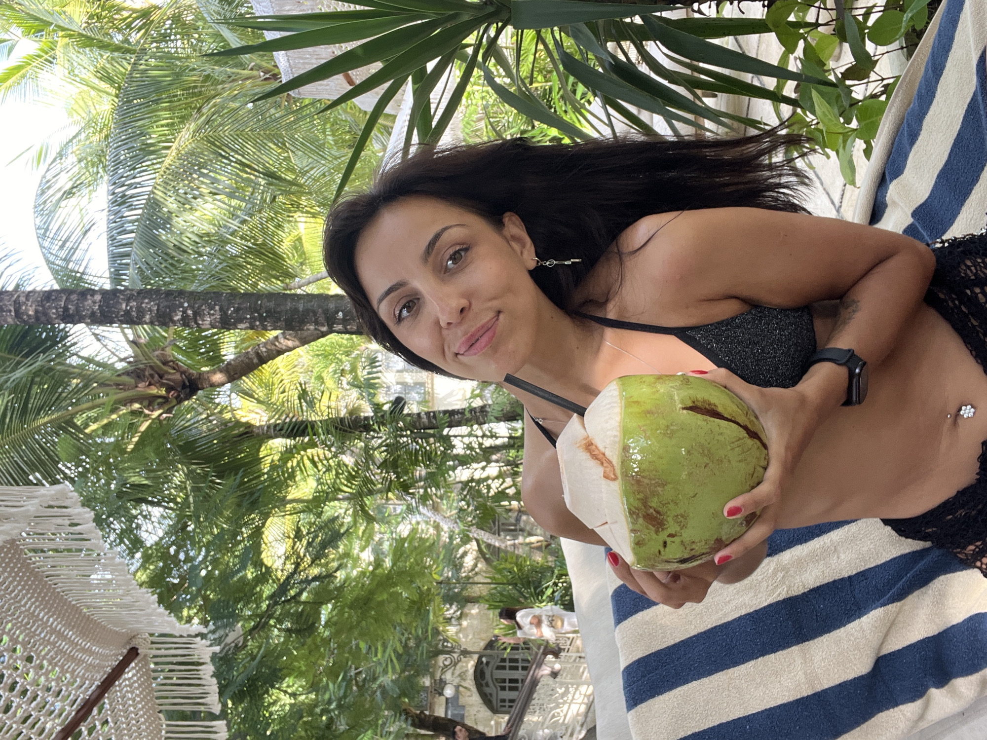 A woman with dark hair is holding a green coconut with a straw in a tropical setting. She is wearing a sparkly black bikini top and has a watch on her left wrist. She is lying on a blue and white striped lounge chair. The background includes palm trees and other tropical foliage. The woman is smiling and looking at the camera. The location is Kuta Selatan, Indonesia.