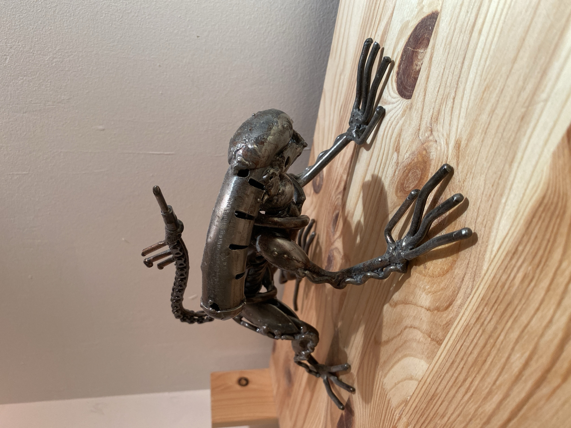 A metallic sculpture of a creature resembling a crab or alien, with segmented limbs and a cylindrical body, is positioned on a wooden surface. The creature is metallic grey and appears to be constructed from various metal parts. Behind the sculpture, a plain, light-colored wall is visible, with a small wooden block on the left side of the frame. The wooden surface has a natural grain pattern. The scene is lit by artificial light, creating shadows on the wooden surface. There are no people or any identifiable Moscow landmarks present.