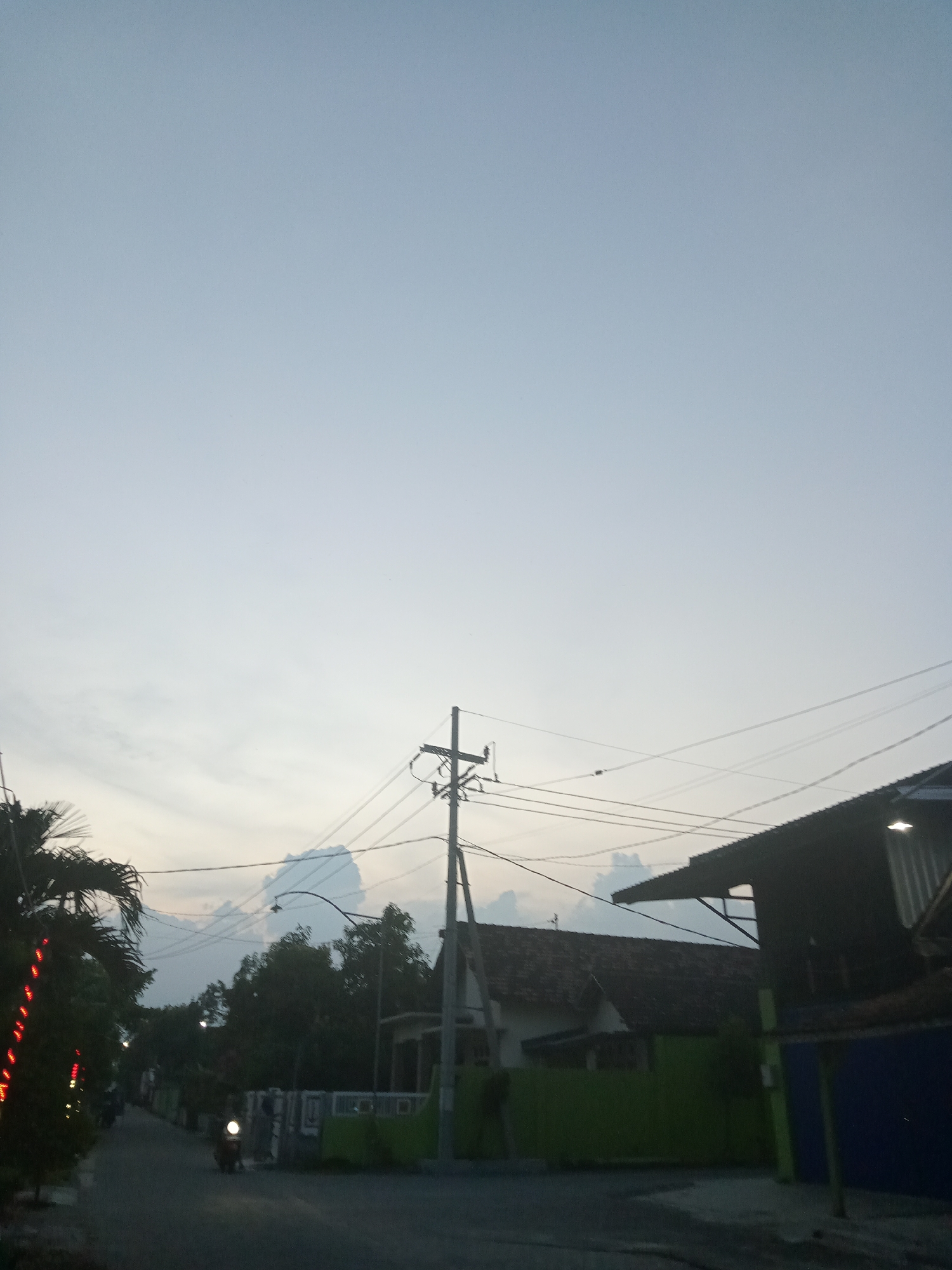 The image depicts a street scene in Bungur, Indonesia, at dusk. A scooter with its headlight on travels down the road. On the left, a palm tree adorned with red lights is visible. Overhead, power lines connect to a utility pole. In the background, houses with green fences and tiled roofs can be seen. The sky is a pale blue, indicating early evening.