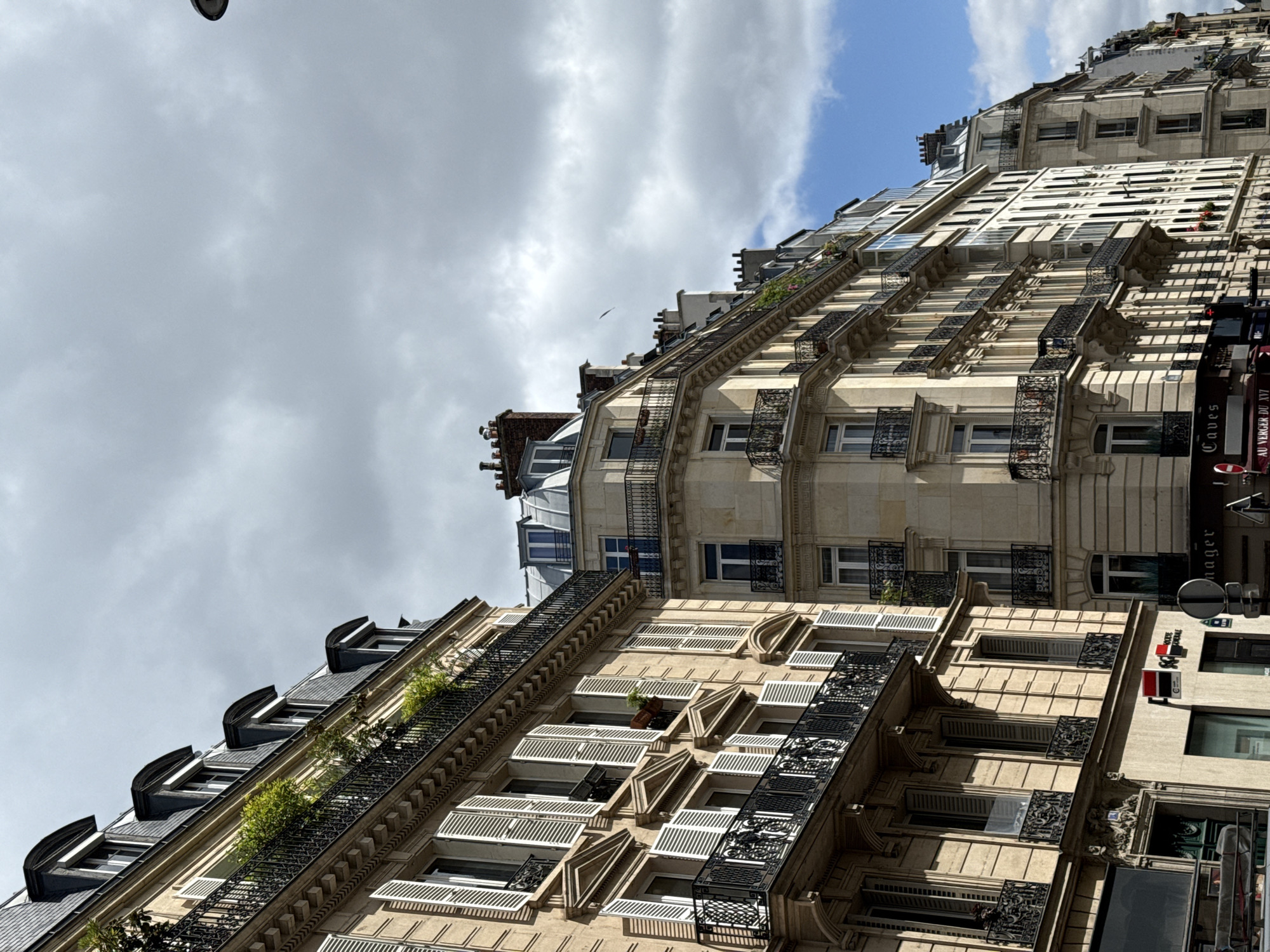 The image shows a street view of typical Parisian buildings in France, captured from a low angle. The buildings have multiple stories, featuring classic architectural details such as wrought iron balconies, window shutters, and ornate facades. The facades are colored in light beige and white. The sky is filled with clouds. A street lamp is visible on the top left corner of the image. Street signs and other building decorations are also visible.