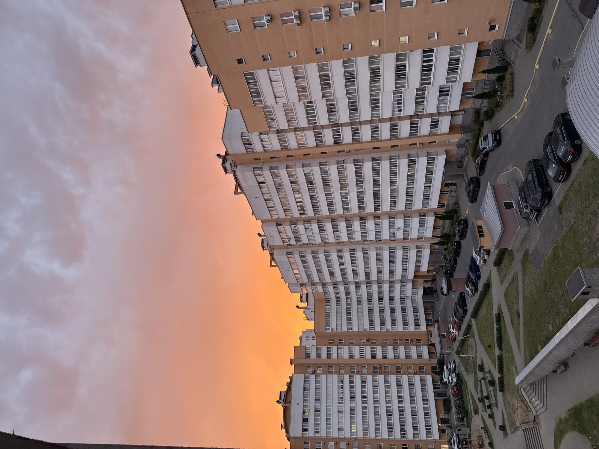 The image is a medium-angle shot of a residential courtyard in Minsk, Belarus, during a vibrant sunset. The sky is a dramatic blend of orange and pink hues, with scattered clouds. Dominating the scene are several multi-story apartment buildings with light-colored facades and orange accents. The buildings are modern, featuring numerous windows and air conditioning units visible on their exteriors.
In the courtyard area, there is a paved road with several parked cars of various makes and colors, predominantly dark sedans and SUVs. A small playground area with swings is visible, along with some landscaping featuring small trees and manicured bushes. A few small, utilitarian buildings are also present within the courtyard. The architecture suggests a post-Soviet era development, common in many large cities. The overall impression is one of urban living during a picturesque evening.