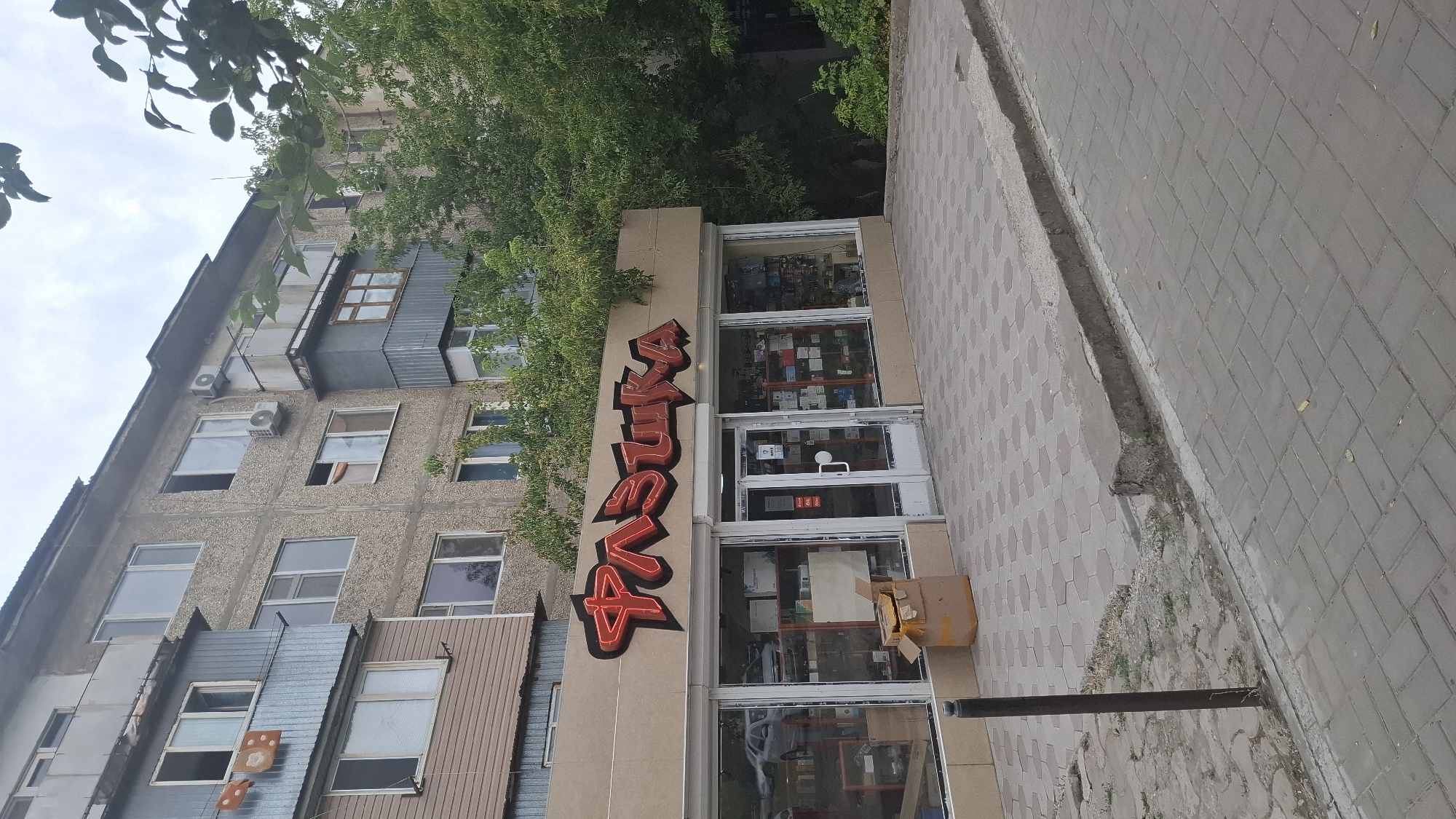 The image depicts the exterior of a store with a sign featuring the stylized red lettering "РАЗУШКА" in the city of Shymkent, Kazakhstan. The store is located on the ground floor of a multi-story apartment building. The storefront has large glass windows displaying various items, and a double glass door serves as the entrance. In front of the store, there is a sidewalk paved with hexagonal and rectangular tiles. A cardboard box is placed on the pavement near the entrance. The building itself shows signs of wear and tear, with some balconies and windows visible. Trees and foliage surround the building, suggesting an outdoor, urban environment. The sky is overcast. There are no people visible in the frame.